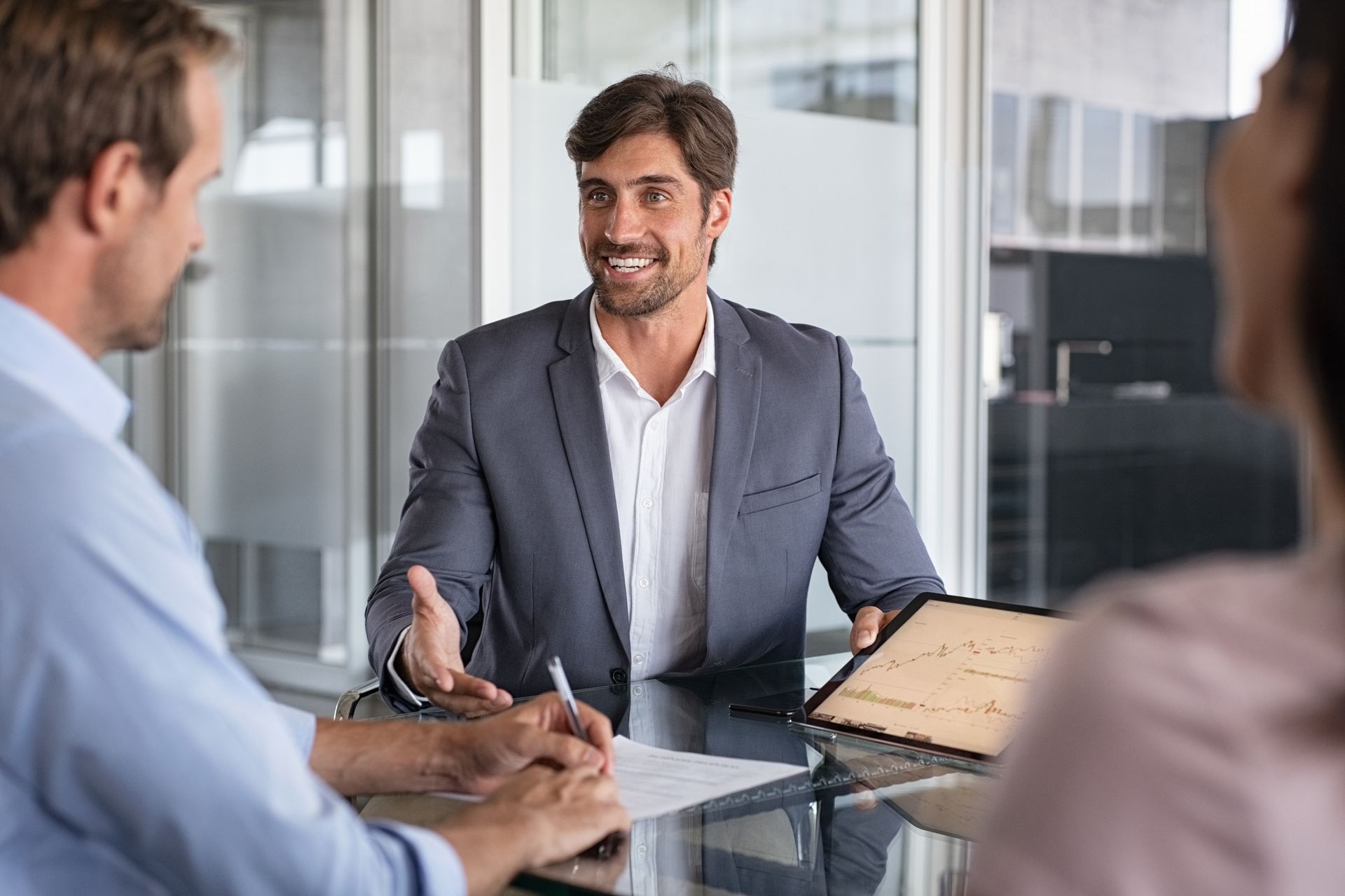 A professional in a suit explains data on a tablet to two colleagues during a business meeting in a modern office.