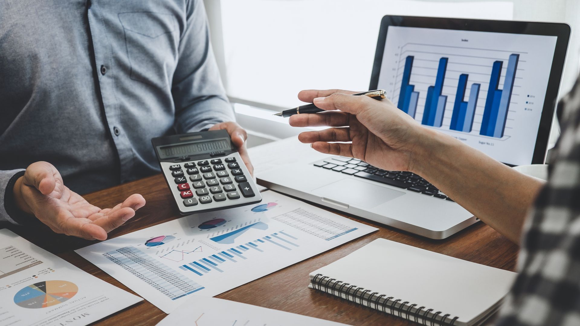 Two people analyze financial charts and graphs on papers and a laptop, using a calculator during a professional meeting.
