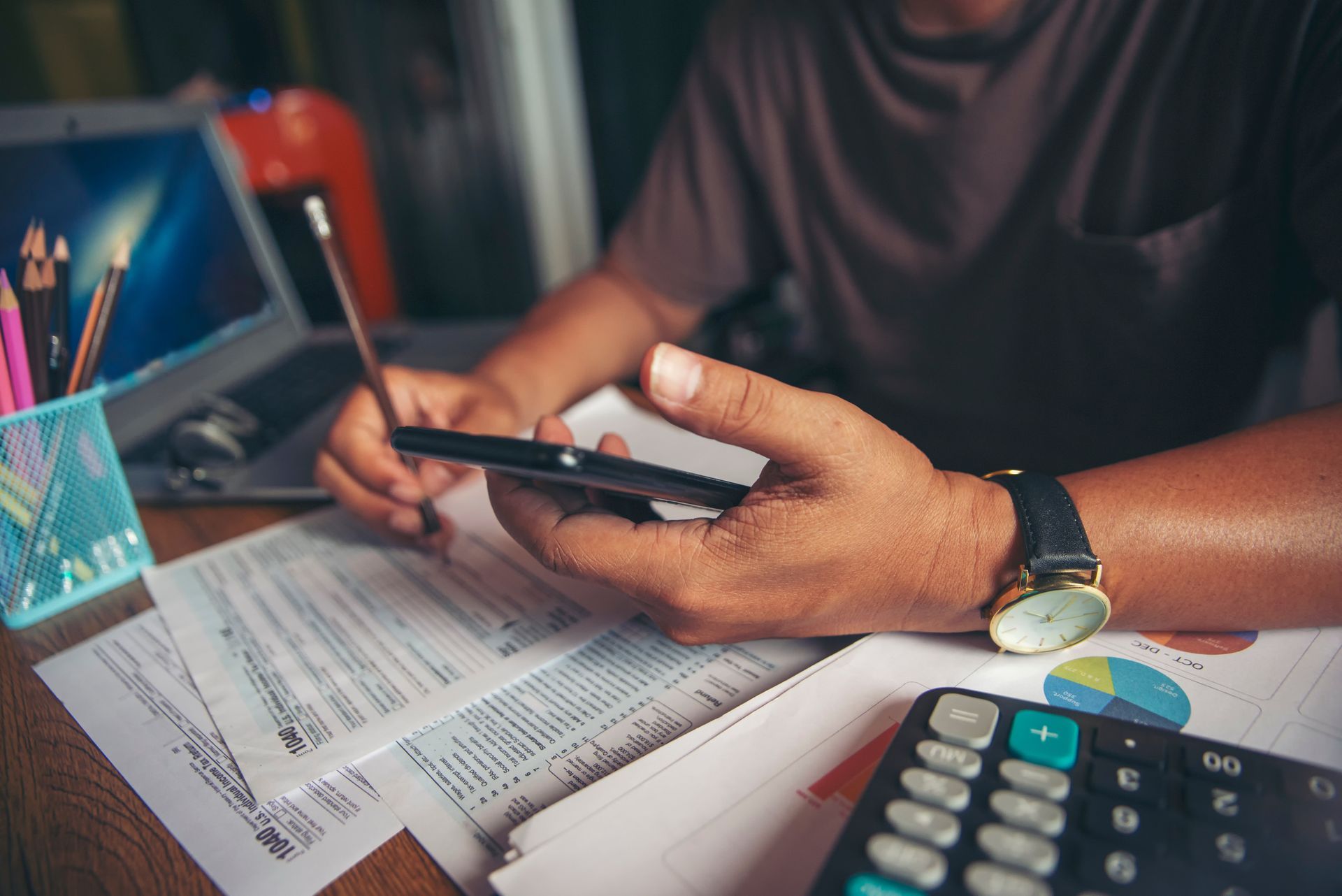 A person works at a desk with financial documents, a calculator, a pencil, and a smartphone.
