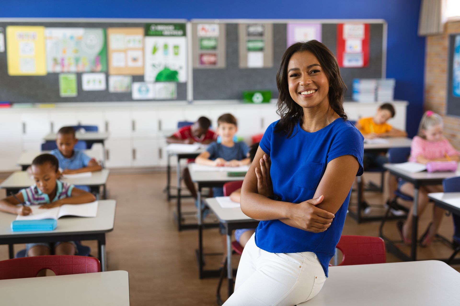 A smiling teacher in a blue shirt leans against a desk in a classroom while students work at their desks in the background.