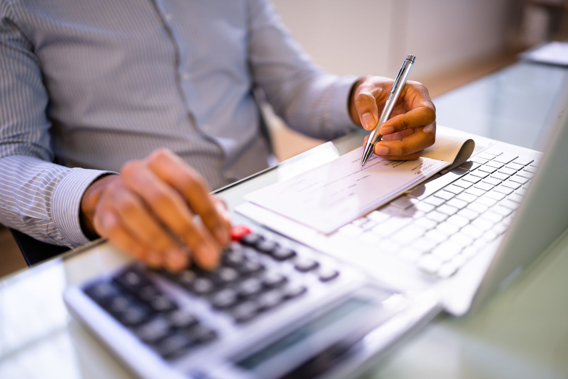 A person writing on a check while using a calculator and a laptop on a desk.