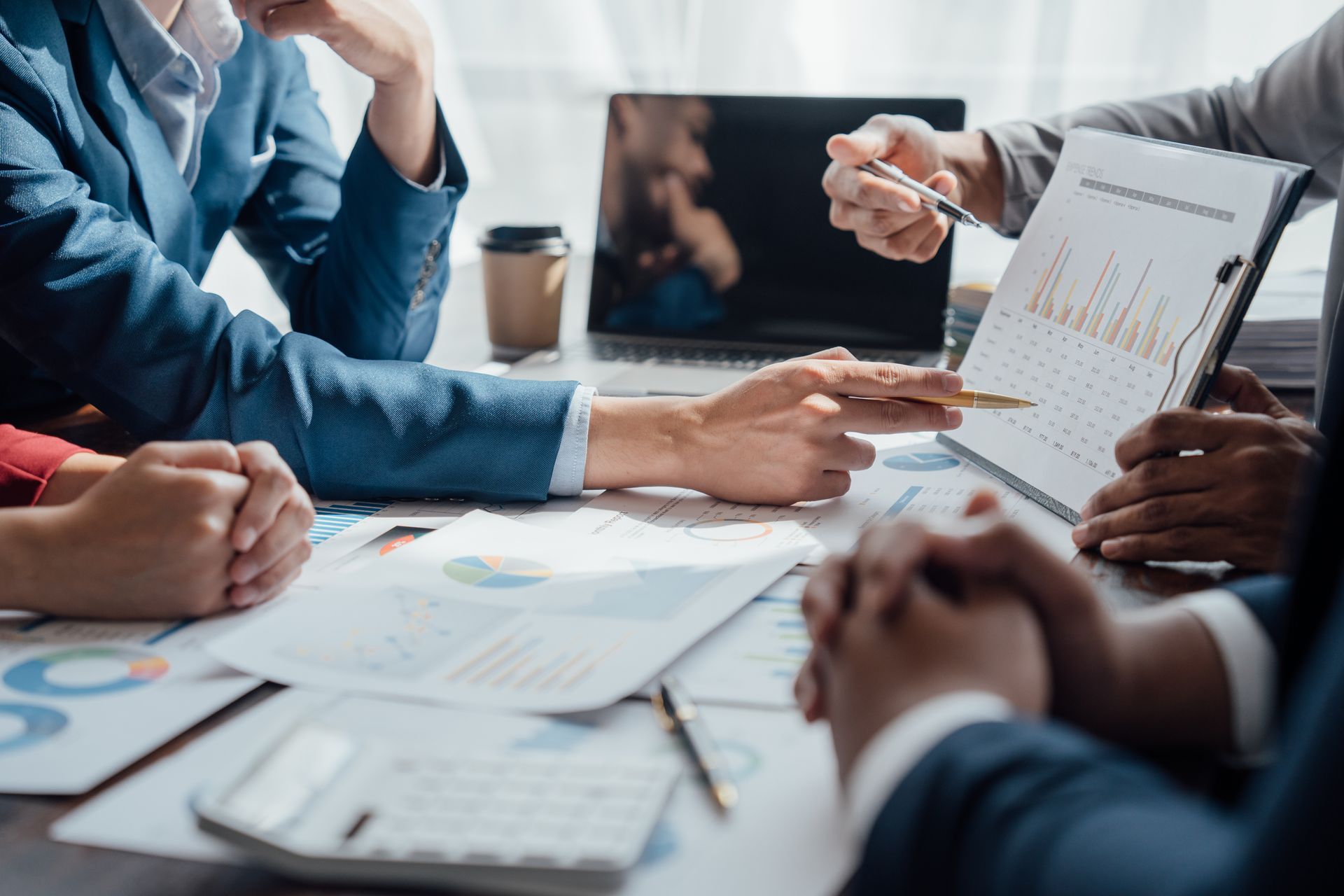 A business team gathers around a table, analyzing charts and data on documents and a laptop during a meeting.