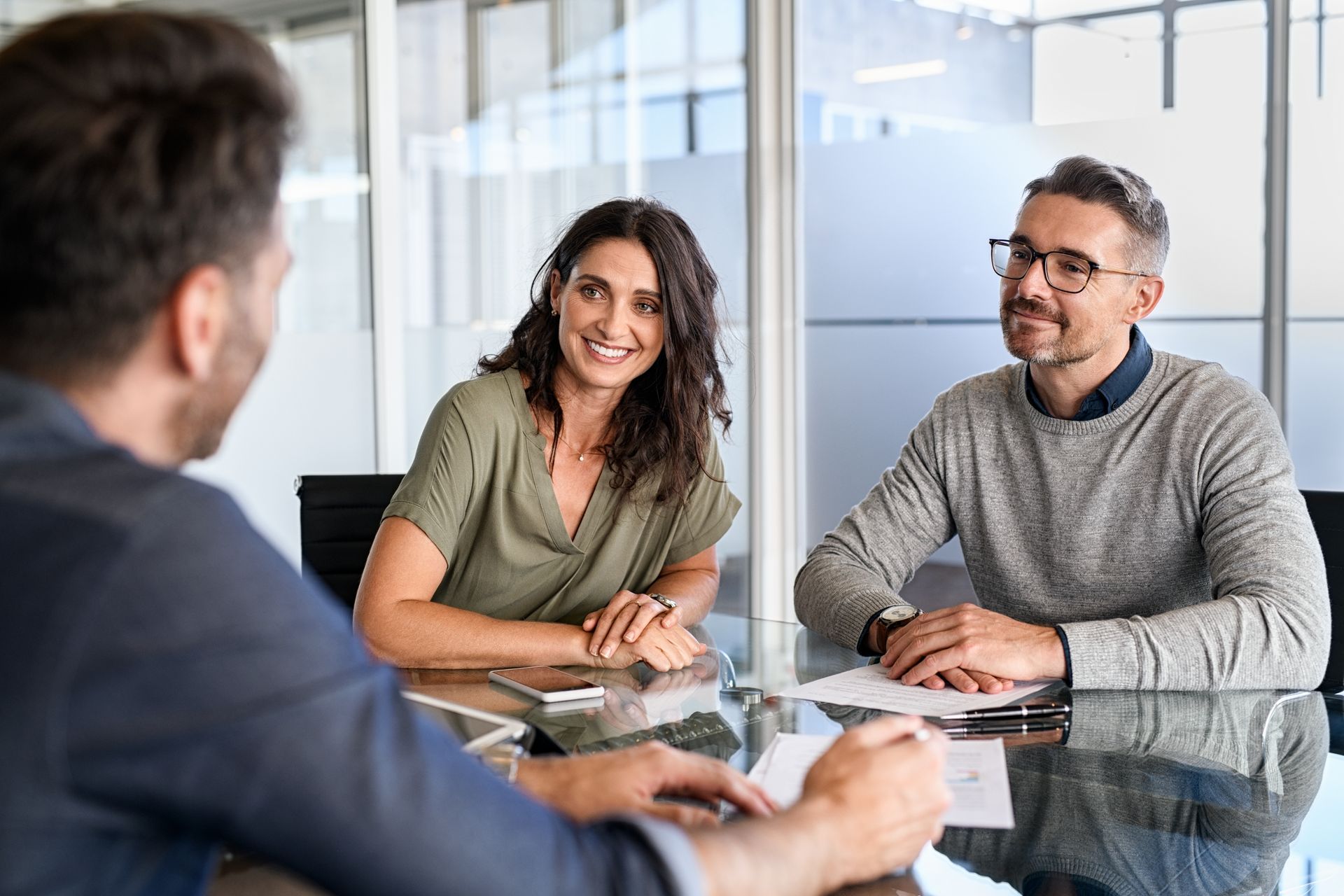 Three professionals sitting at a glass conference table in a bright office, engaging in a collaborative discussion.