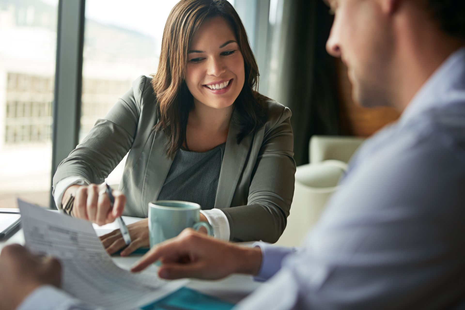Two people sit at a table reviewing documents together in a sunlit office, smiling and engaged in professional discussion.