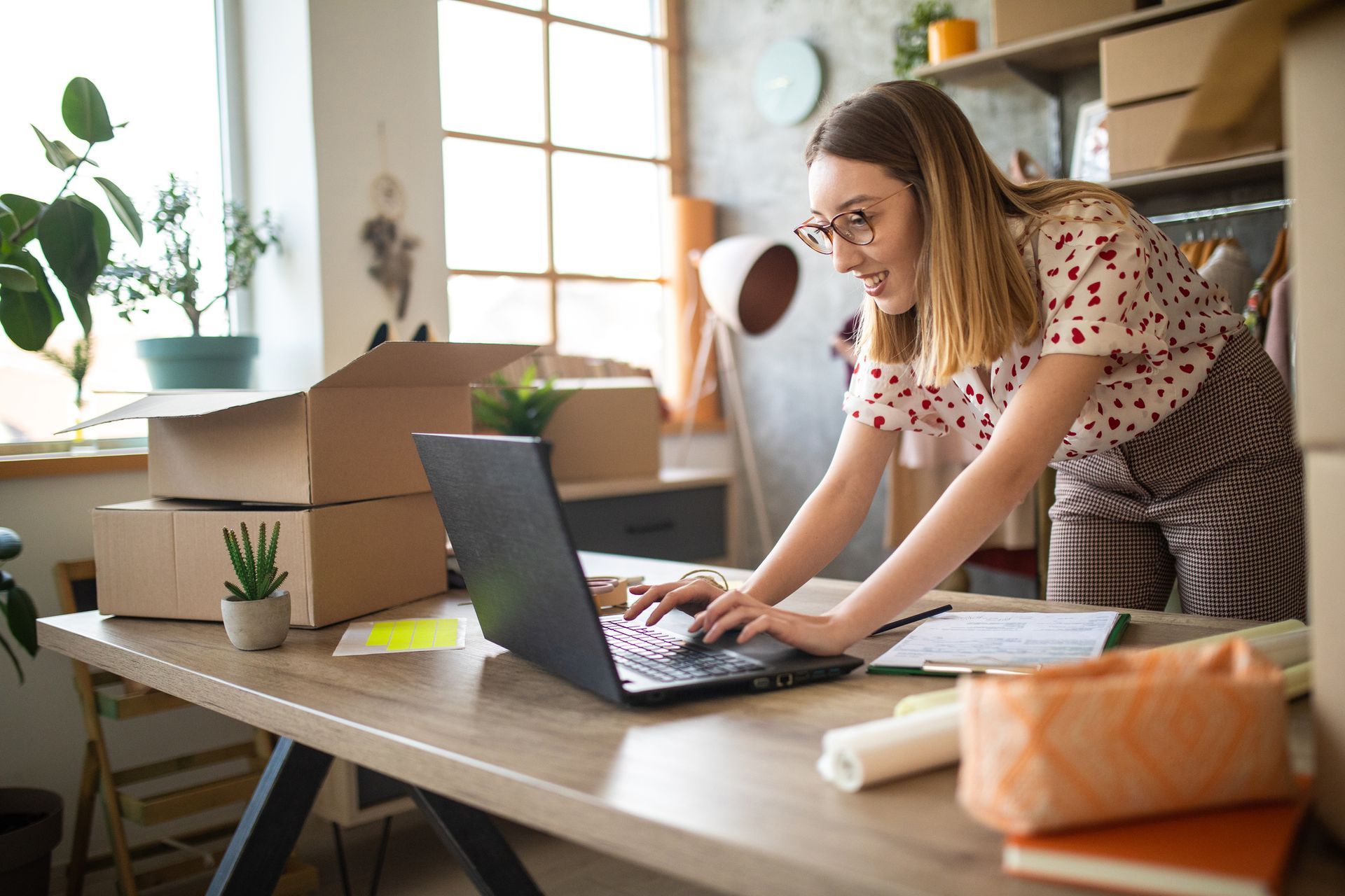 A person wearing glasses works at a laptop on a desk filled with shipping boxes in a well-lit home office.