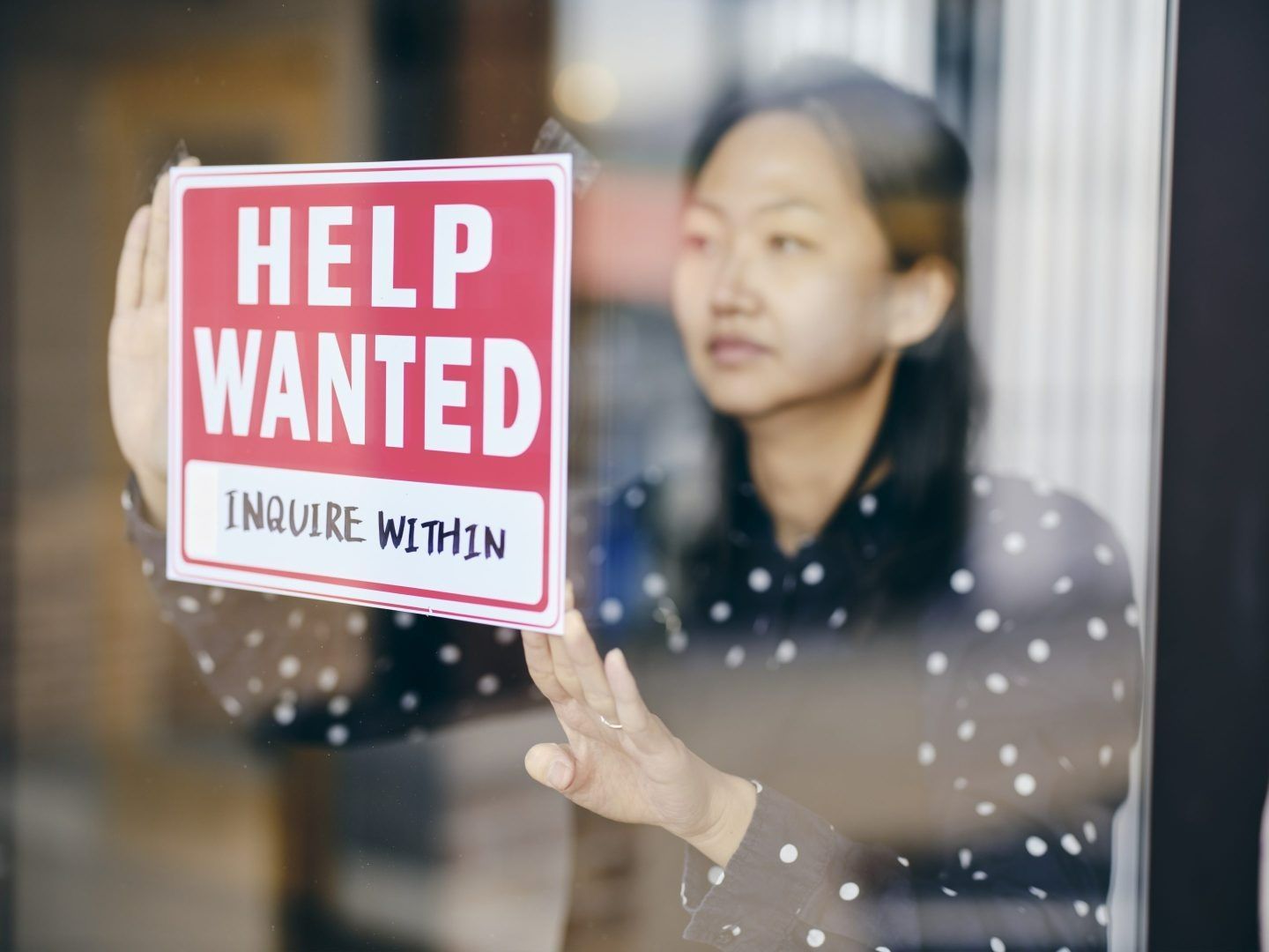 A person in a polka-dot shirt puts up a red 'HELP WANTED' sign on a glass window.