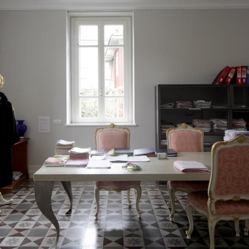 Ufficio con tavolo, sedie, finestra e libreria. Documenti sul tavolo e sugli scaffali. Pavimento piastrellato nero e grigio.