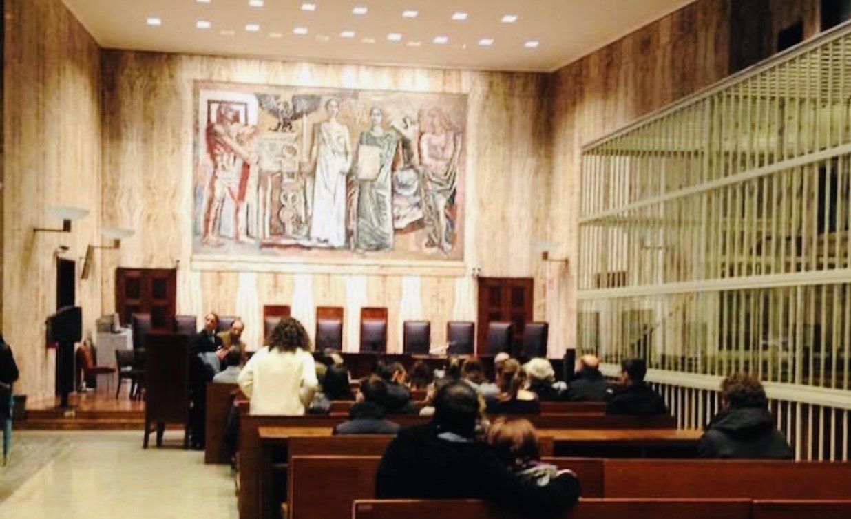 Vista interna di un'aula di tribunale, con persone sedute, un murale sulla parete e una grande gabbia di metallo sulla destra.