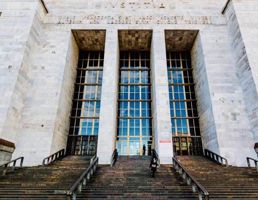 Grande edificio in pietra con tre alte porte in vetro, gradini e persone che entrano.