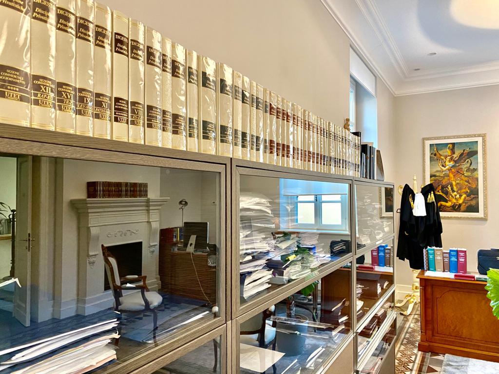 Scaffali pieni di libri di giurisprudenza sopra una scrivania in un ufficio con camino e quadri alle pareti.