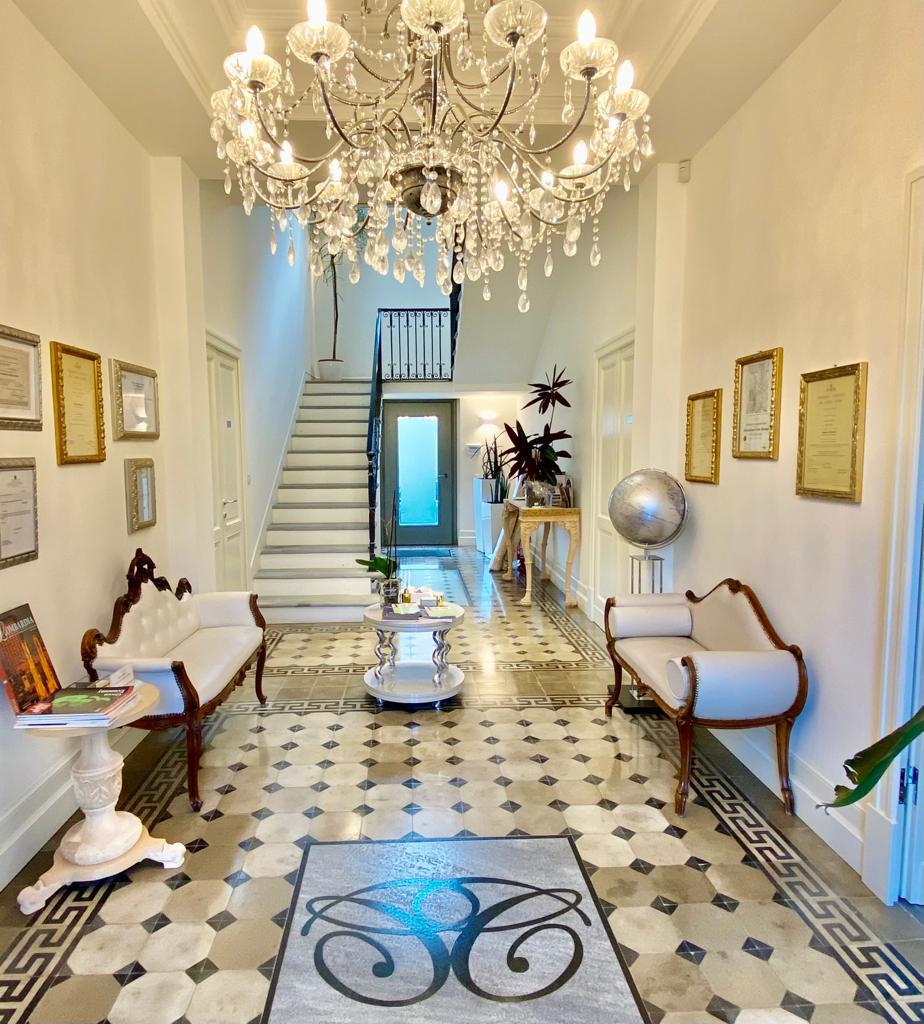 Elegante corridoio con lampadario di cristallo, scala, mobili bianchi, quadri incorniciati e pavimento in piastrelle a motivi.