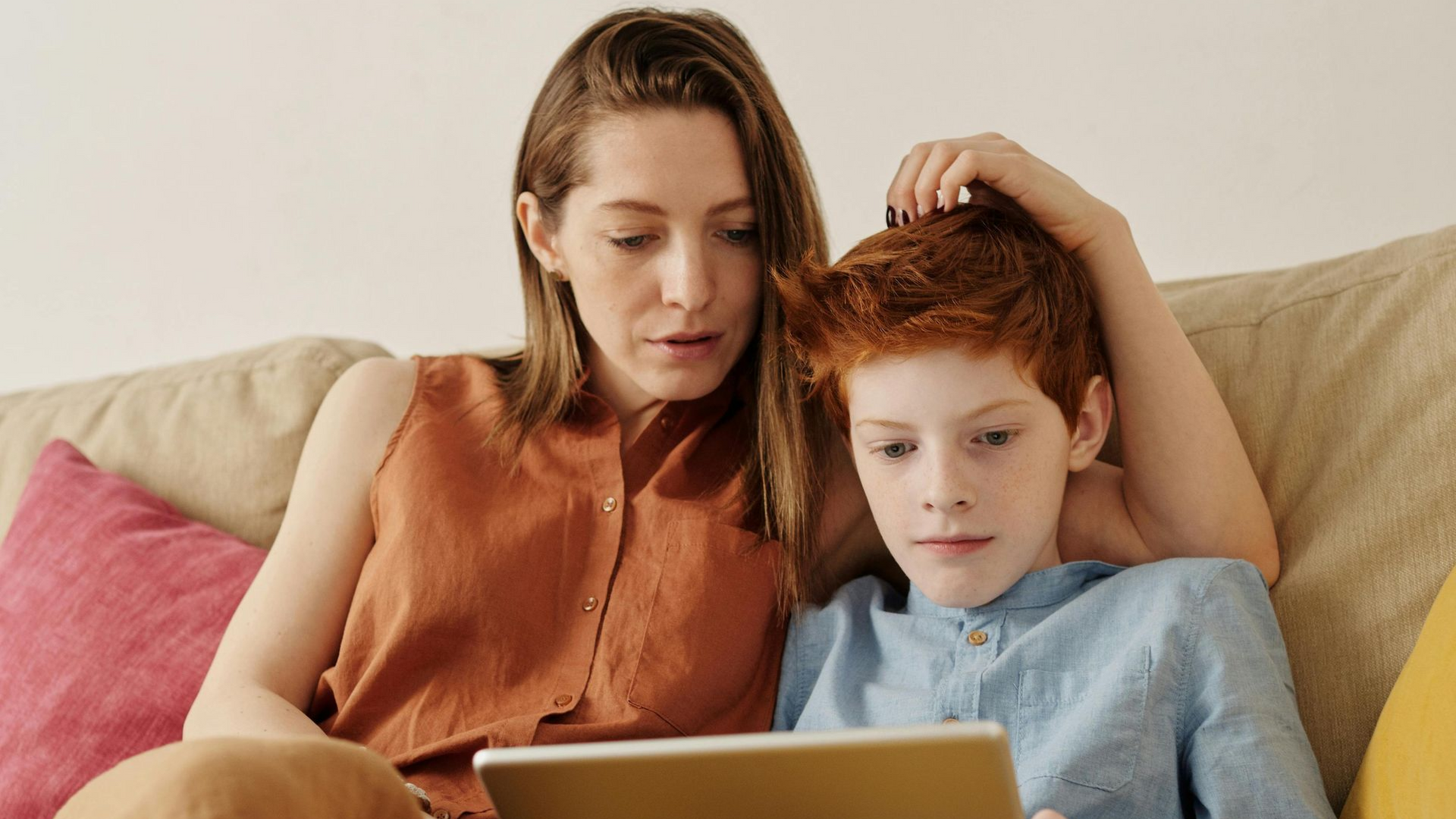 Woman and child looking at a tablet together while seated on a couch. The woman is wearing a brown top and the child has red hair. PARENTAL CONTROLS