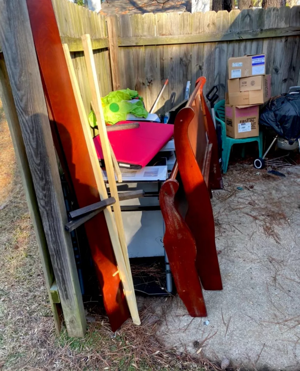 Wooden furniture pieces and boxes stacked next to a wooden fence and a trash can in a backyard setting.