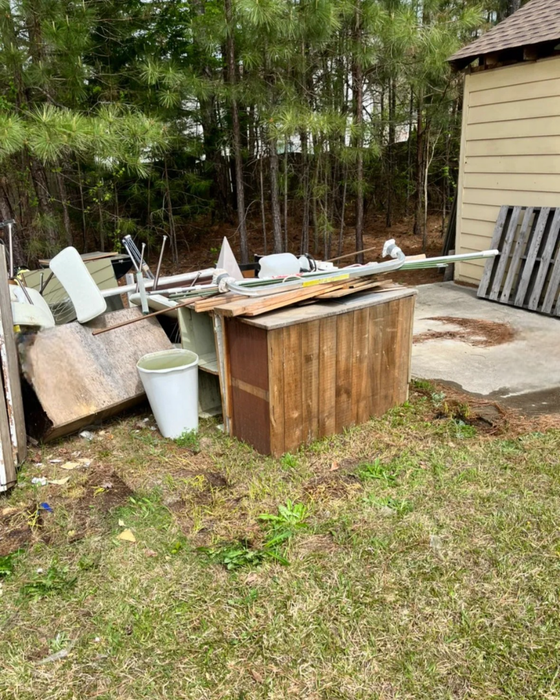 Backyard cluttered with assorted debris, including a wooden structure, pallets, and white containers, near a building and trees.