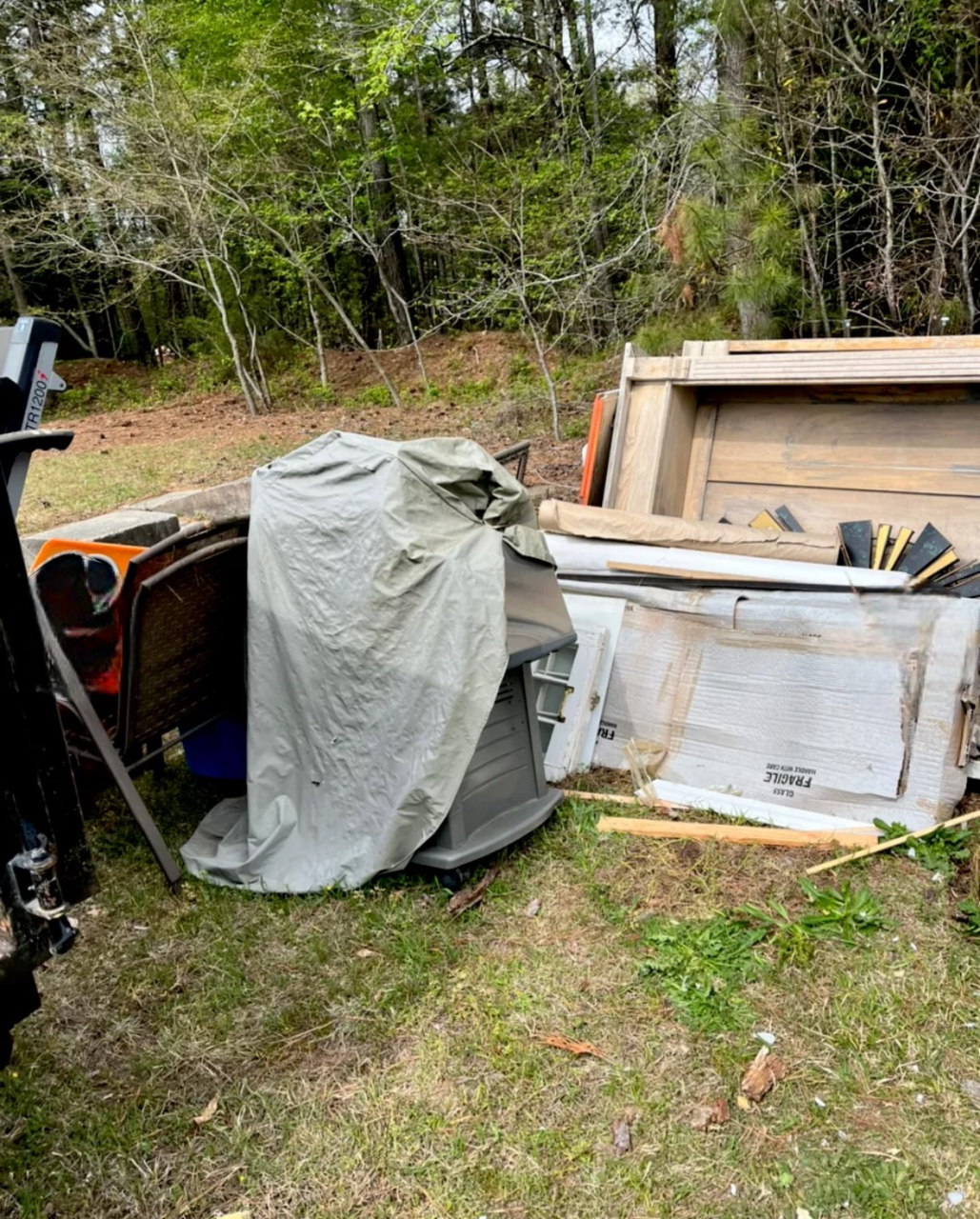 Outdoor scene with tarp-covered object, possible appliance. Old car part and other items, including lumber, are strewn nearby.