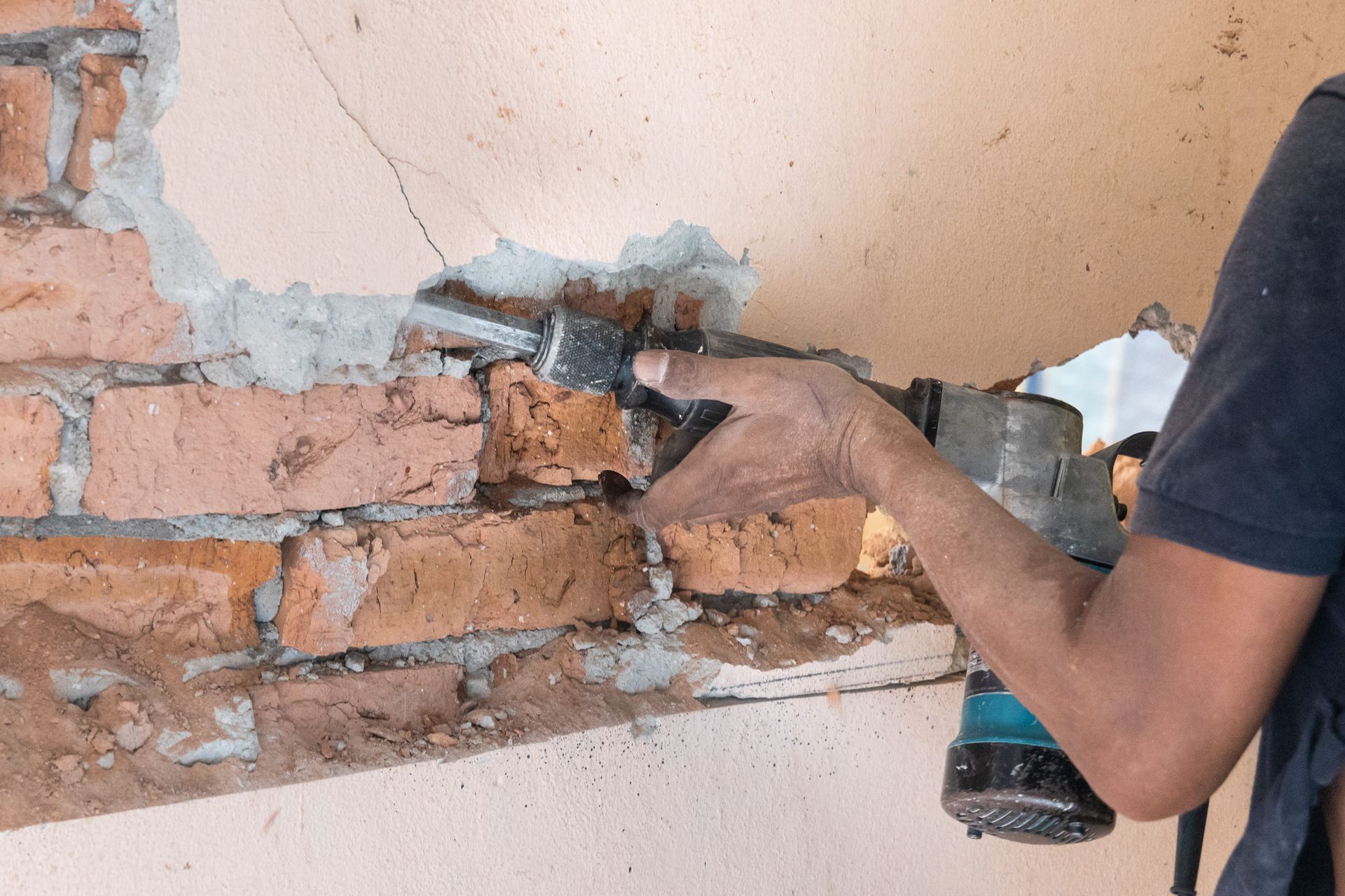 A person using a power tool to remove bricks from a wall. The wall is brick and mortar and partially covered with pink plaster.