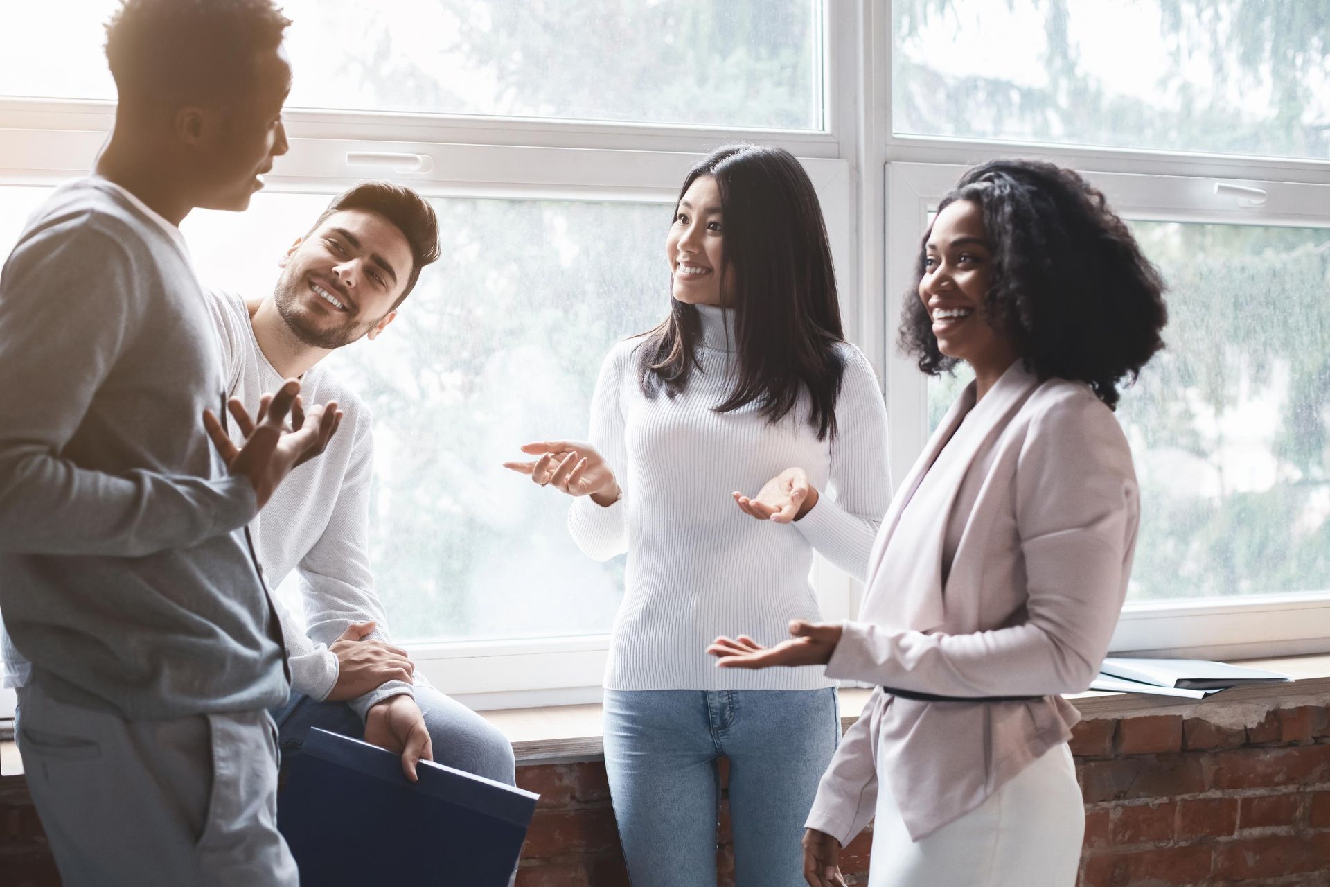Group of diverse young adults chatting near a window, smiling and gesturing in a bright room.
