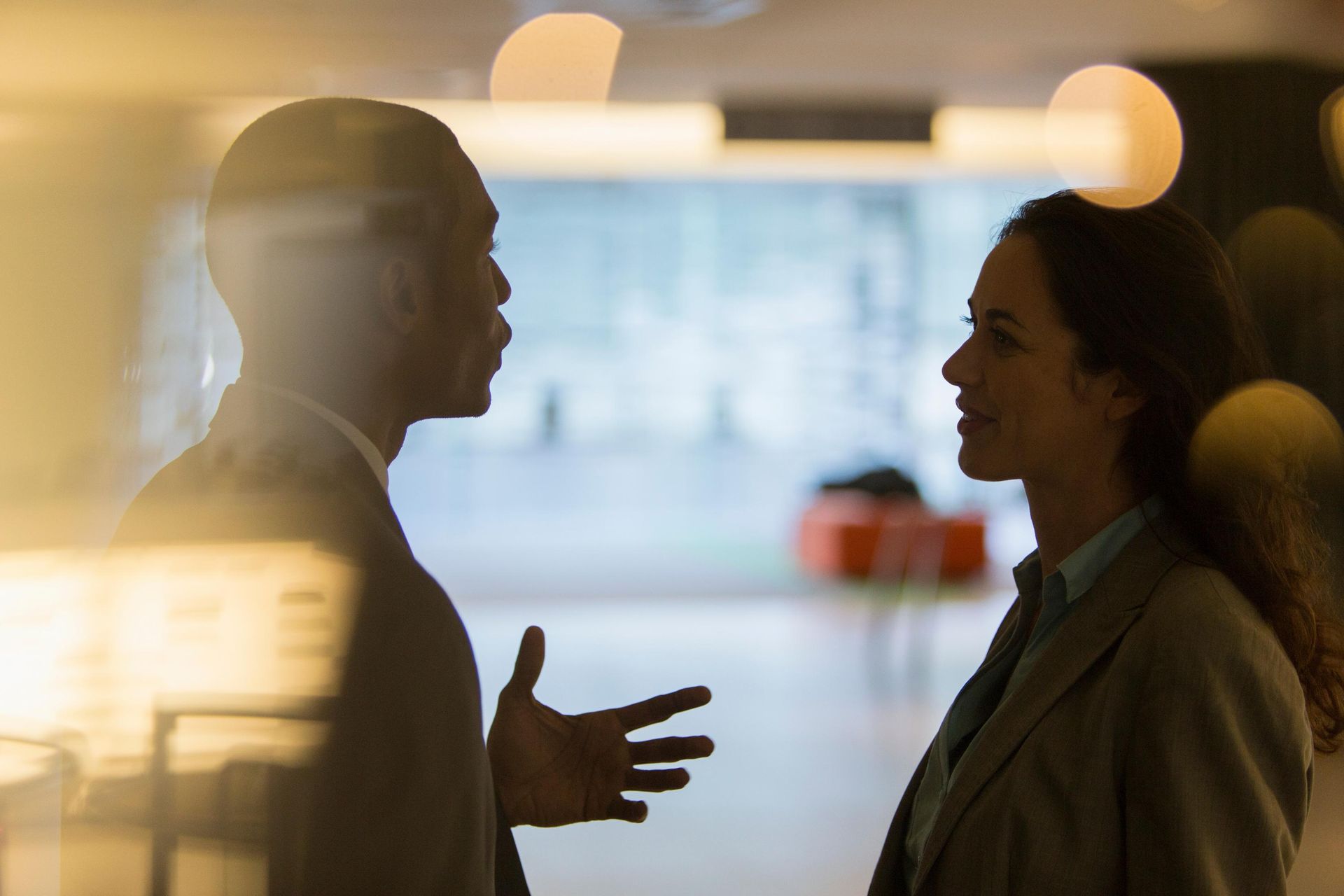 Man and woman in business attire converse in office setting.