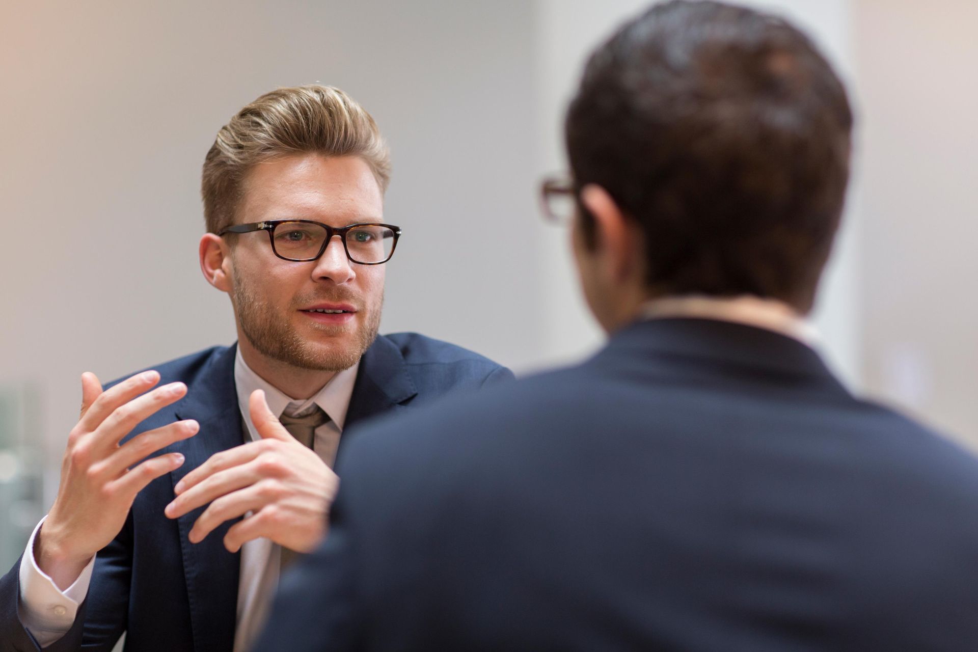 Man in glasses gestures, speaking to person in suit, possibly in an office.