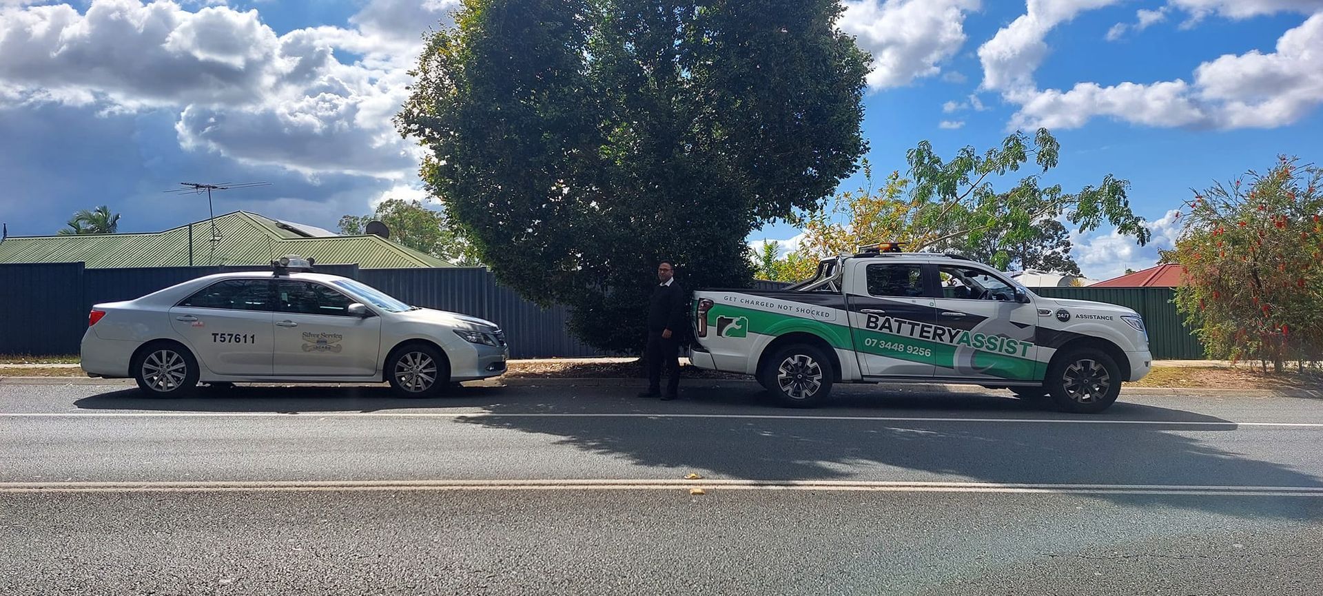 A white car and a pickup truck are parked on a street. A person stands between them, under a large tree — Battery Assist Brisbane in Narangba, QLD