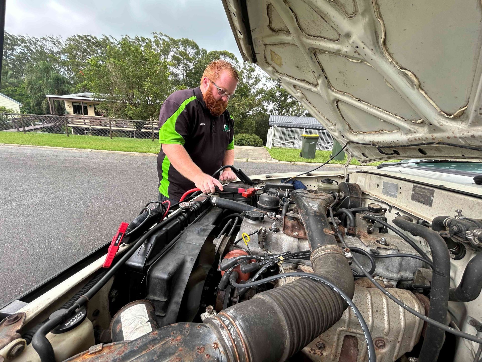 Mechanic working on the engine of a car with the hood open. Wearing a green and black shirt, using jumper cables — Battery Assist Brisbane in Caboolture, QLD