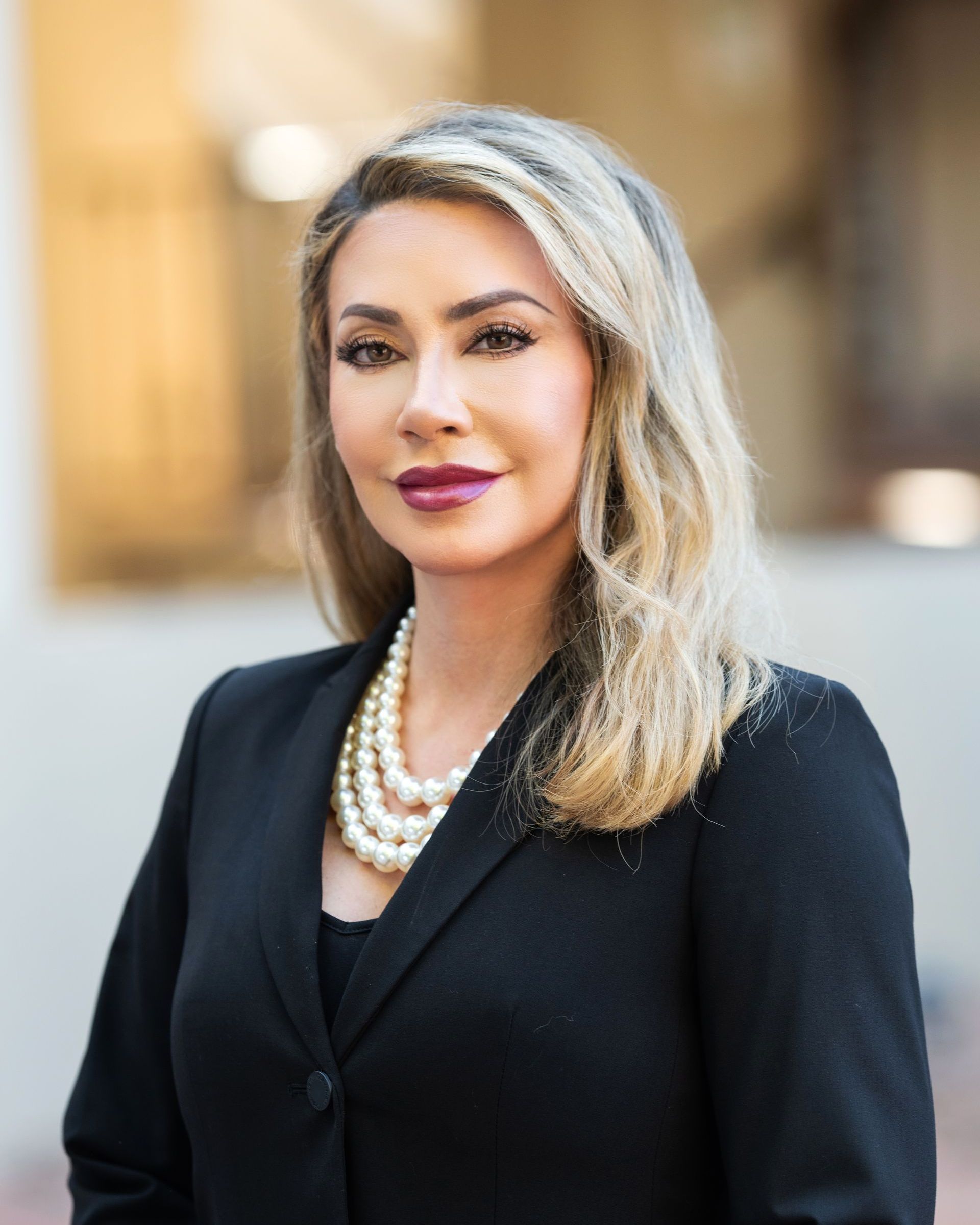 A woman wearing a black jacket and a pearl necklace is standing in front of a building.