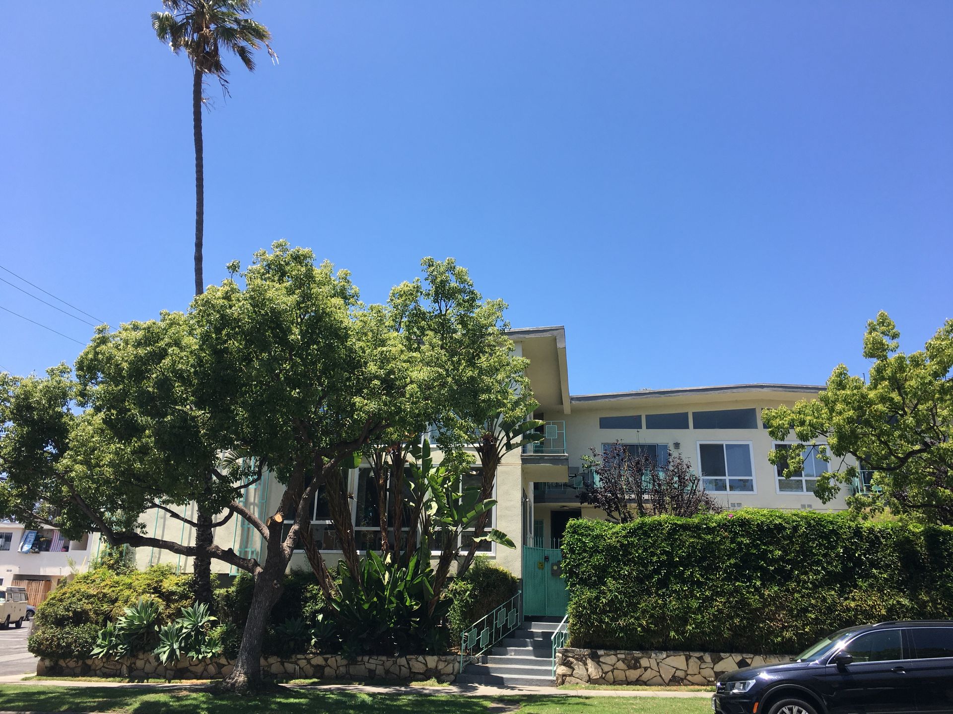 Yellow apartment building with green trees under a blue sky, black car in foreground.