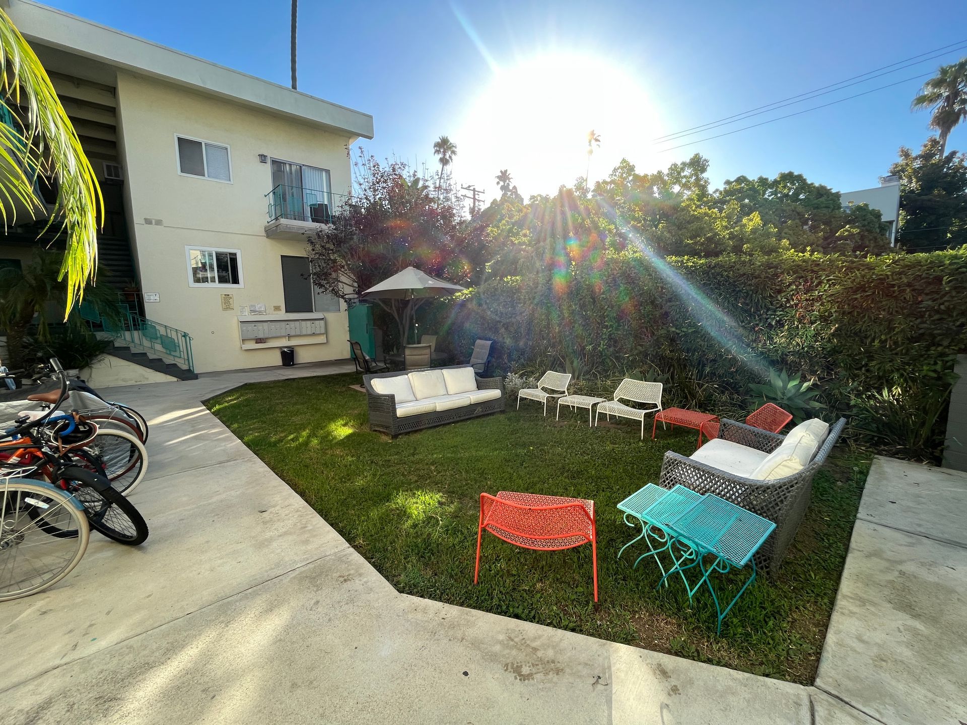 Apartment building with a grassy courtyard, patio furniture, and bicycles on the left, bright sunshine.