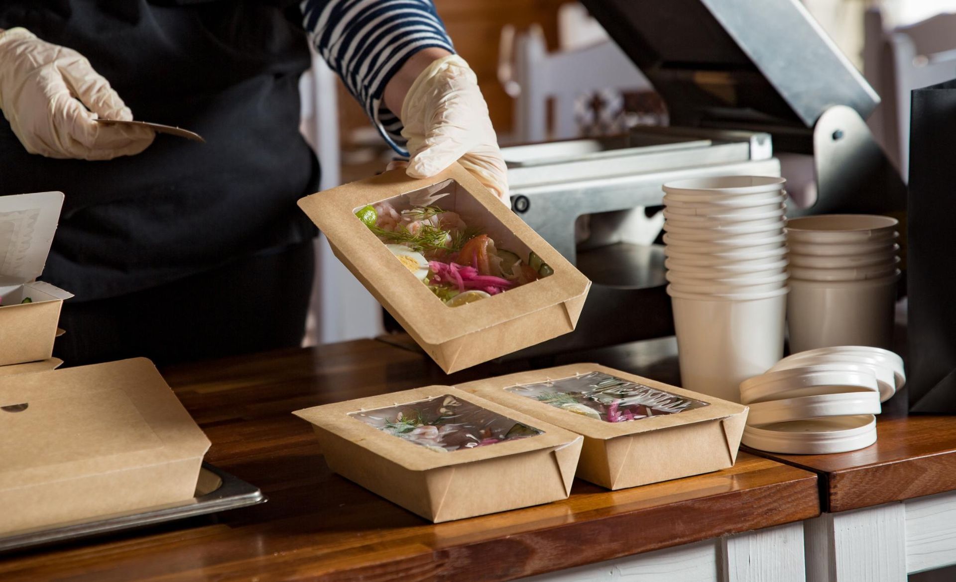 Person wearing gloves holding a takeout food container near other prepared containers on a counter.