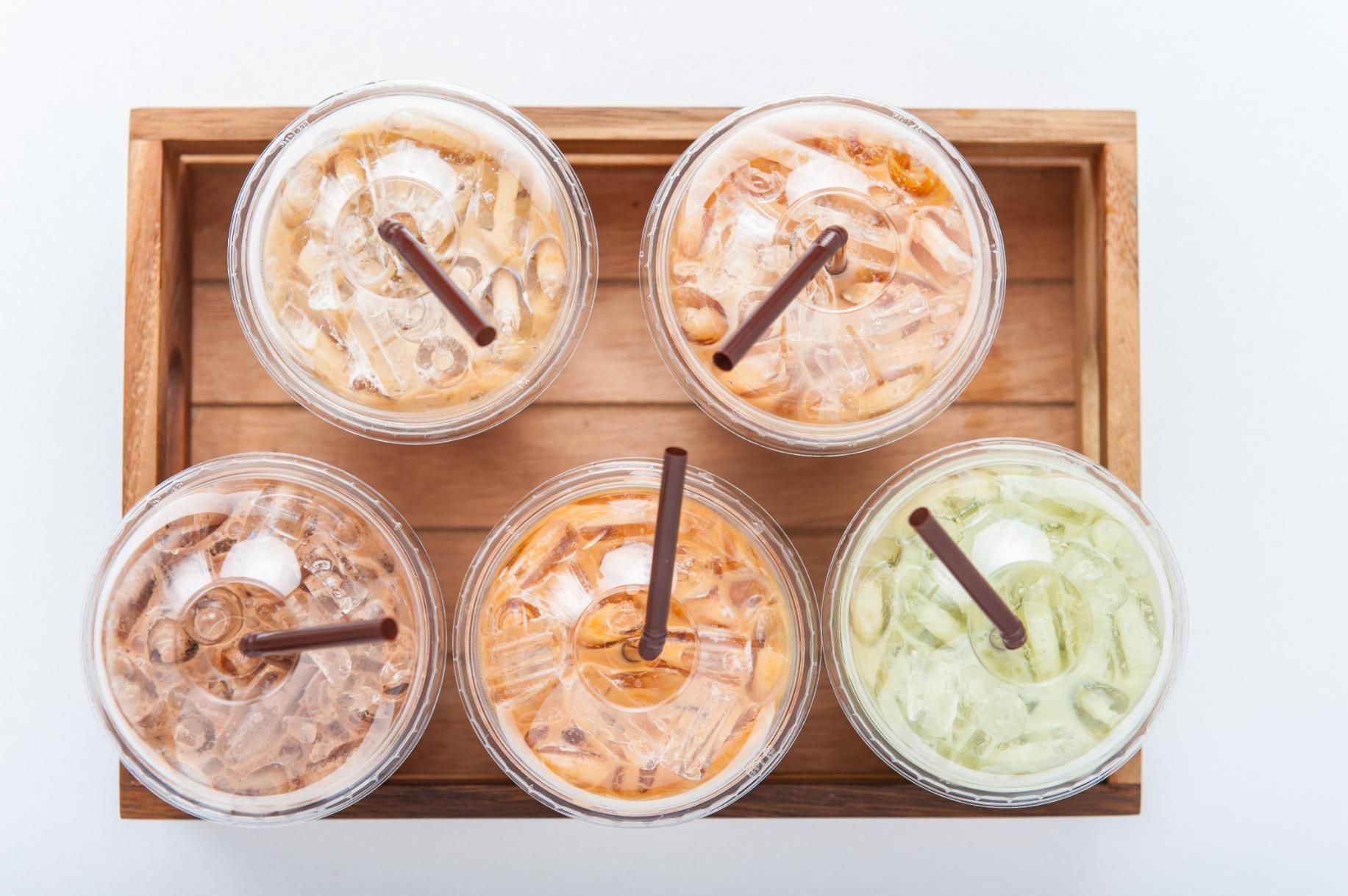Five iced drinks in clear cups with straws in a wooden tray.