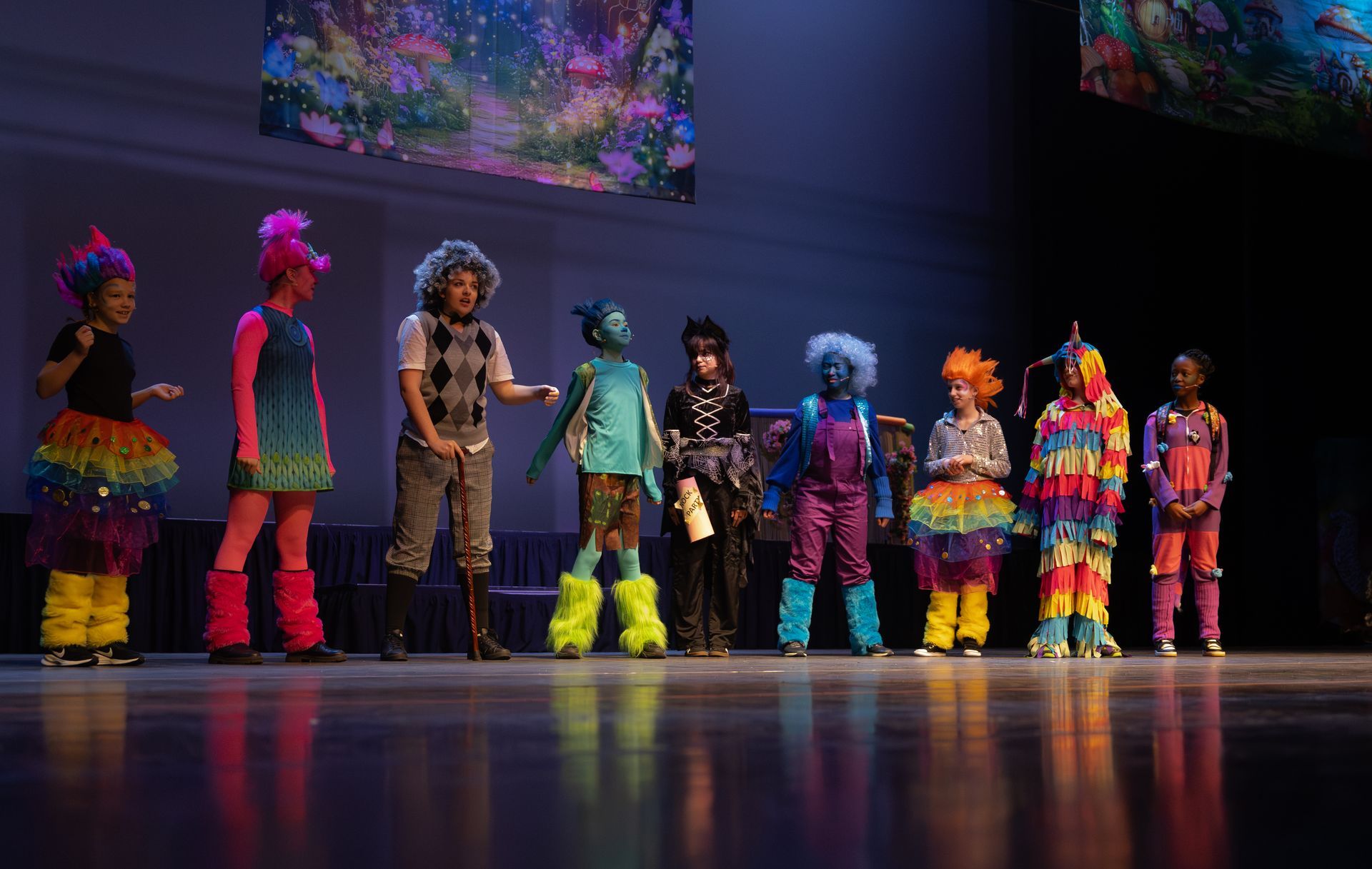Een groep artiesten in kostuums, met kleurrijke pruiken en outfits, staat op een podium. De achtergronden zijn versierd met fantasierijke kunstwerken.