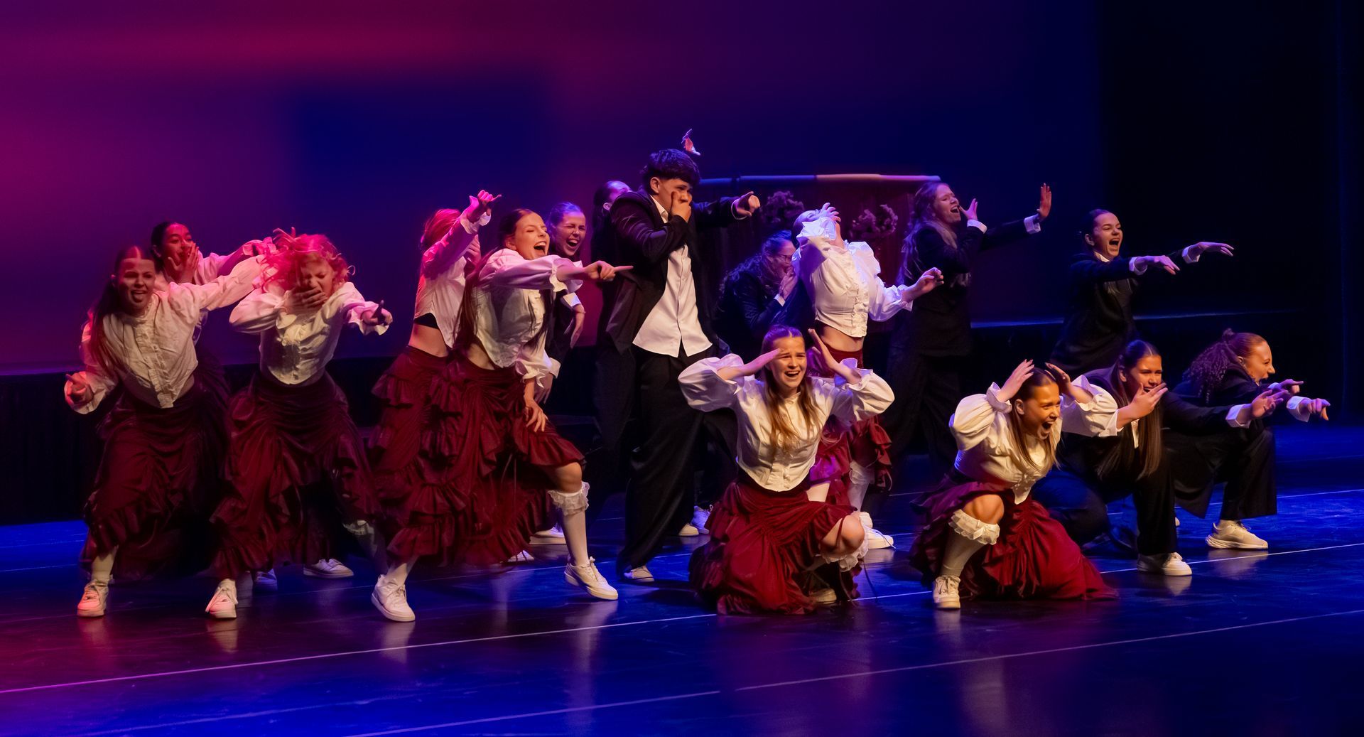 Een groep dansers staat op het podium, sommigen knielen, gekleed in witte blouses en rode rokken. Een man in het midden zingt.
