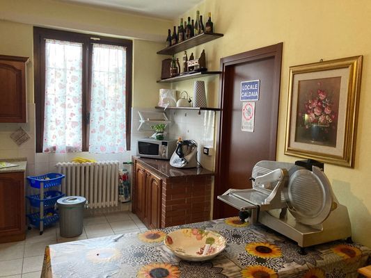 Cucina con finestra, mobiletto e tavolo coperto da una tovaglia a forma di girasole. Affettatrice sul tavolo.