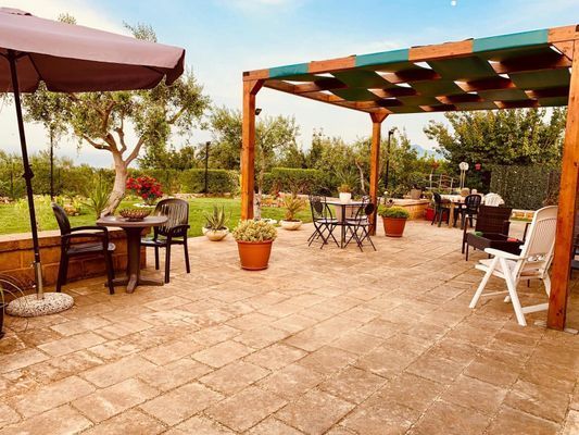 Patio con tavoli e sedie sotto un pergolato e un ombrellone, piante e un giardino.