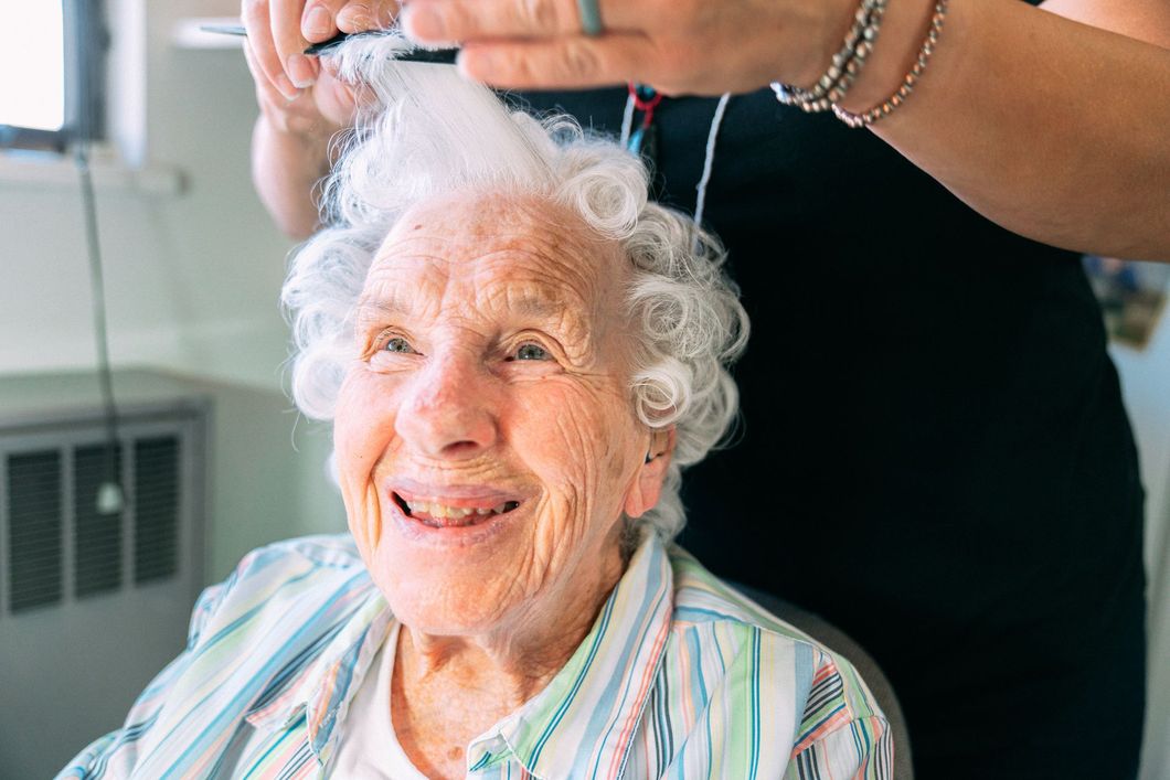 Donna sorridente che si taglia i capelli, in casa. Una persona le tiene le forbici sopra la testa.