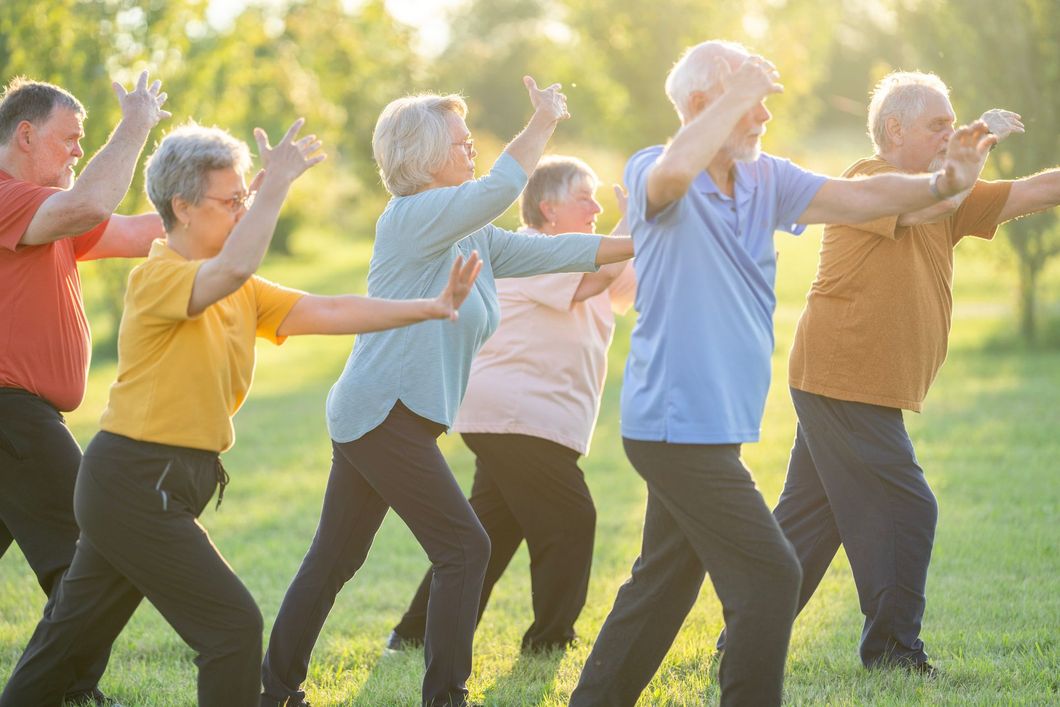 Gruppo di persone in un parco che praticano tai chi. Braccia alzate, sfondo illuminato dal sole.