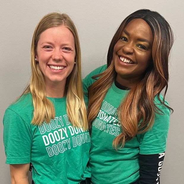 Two smiling people in green T-shirts posing indoors against a plain wall