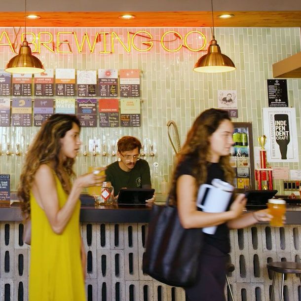 Brewery taproom counter with hanging lights, menu boards, and two blurred customers passing by in the foreground