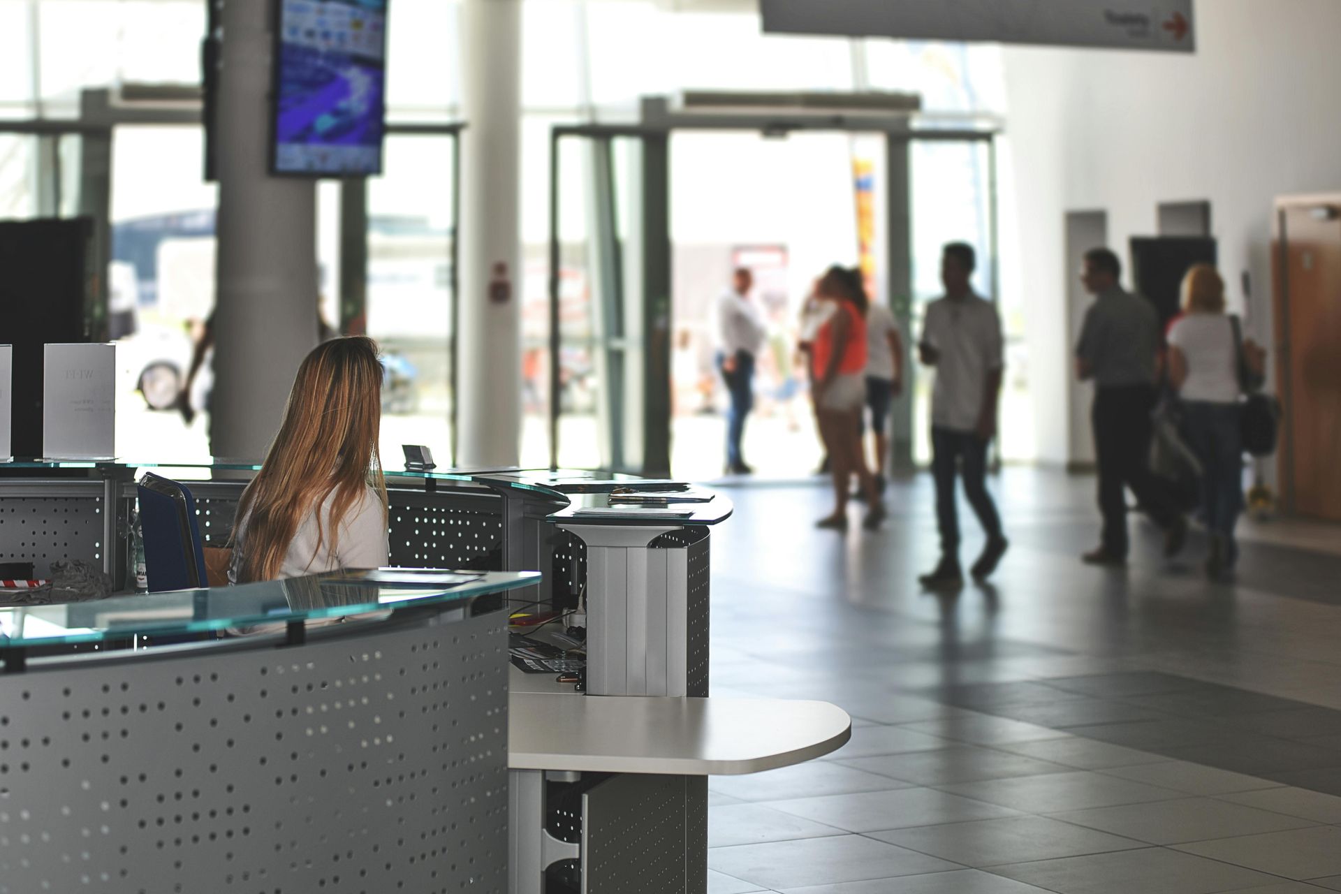 A woman is sitting at a desk in a lobby with people walking around.