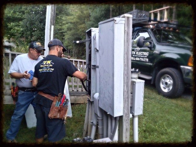 Two electricians wearing tool belts work on an outdoor electrical service panel near a utility truck.