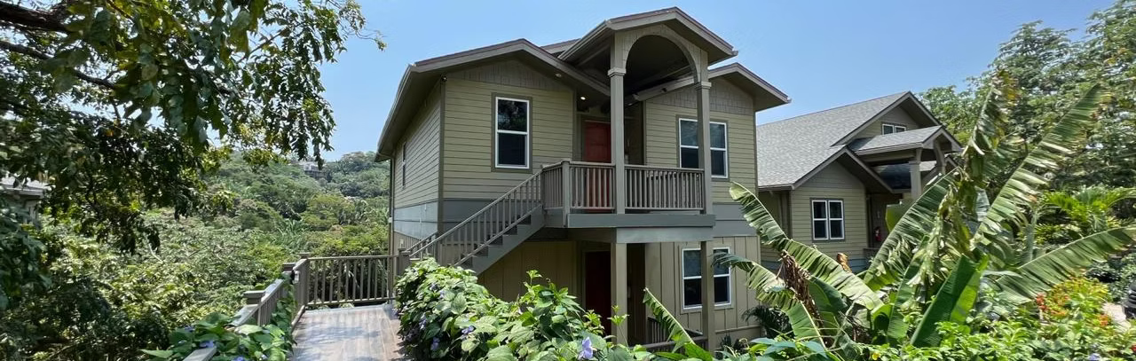 Casa con escalera de madera y balcón, rodeada de exuberante vegetación. Cielo azul de fondo.