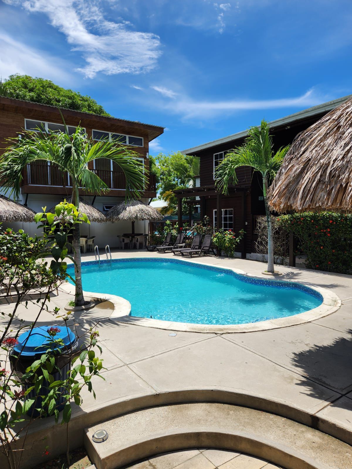 Piscina rodeada de edificios tropicales y árboles bajo un cielo azul.