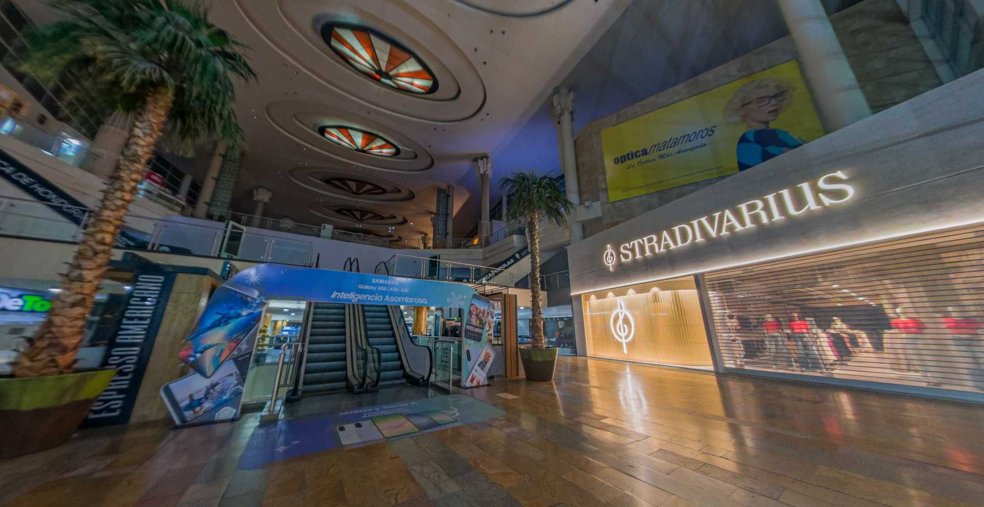 Interior de un centro comercial con una tienda Stradivarius, escaleras mecánicas y palmeras.