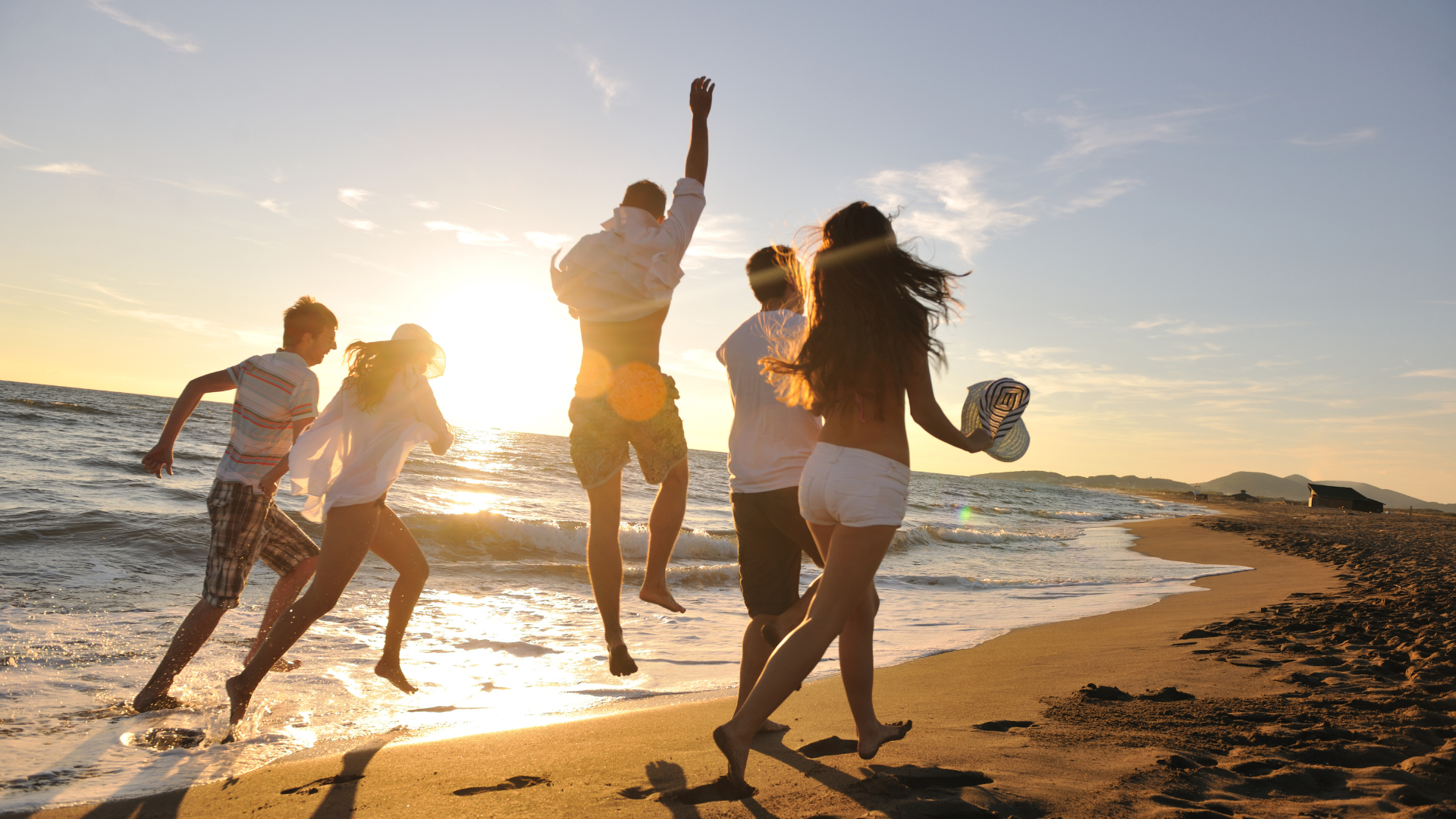 Group of friends running on a beach at sunset, one person jumping.