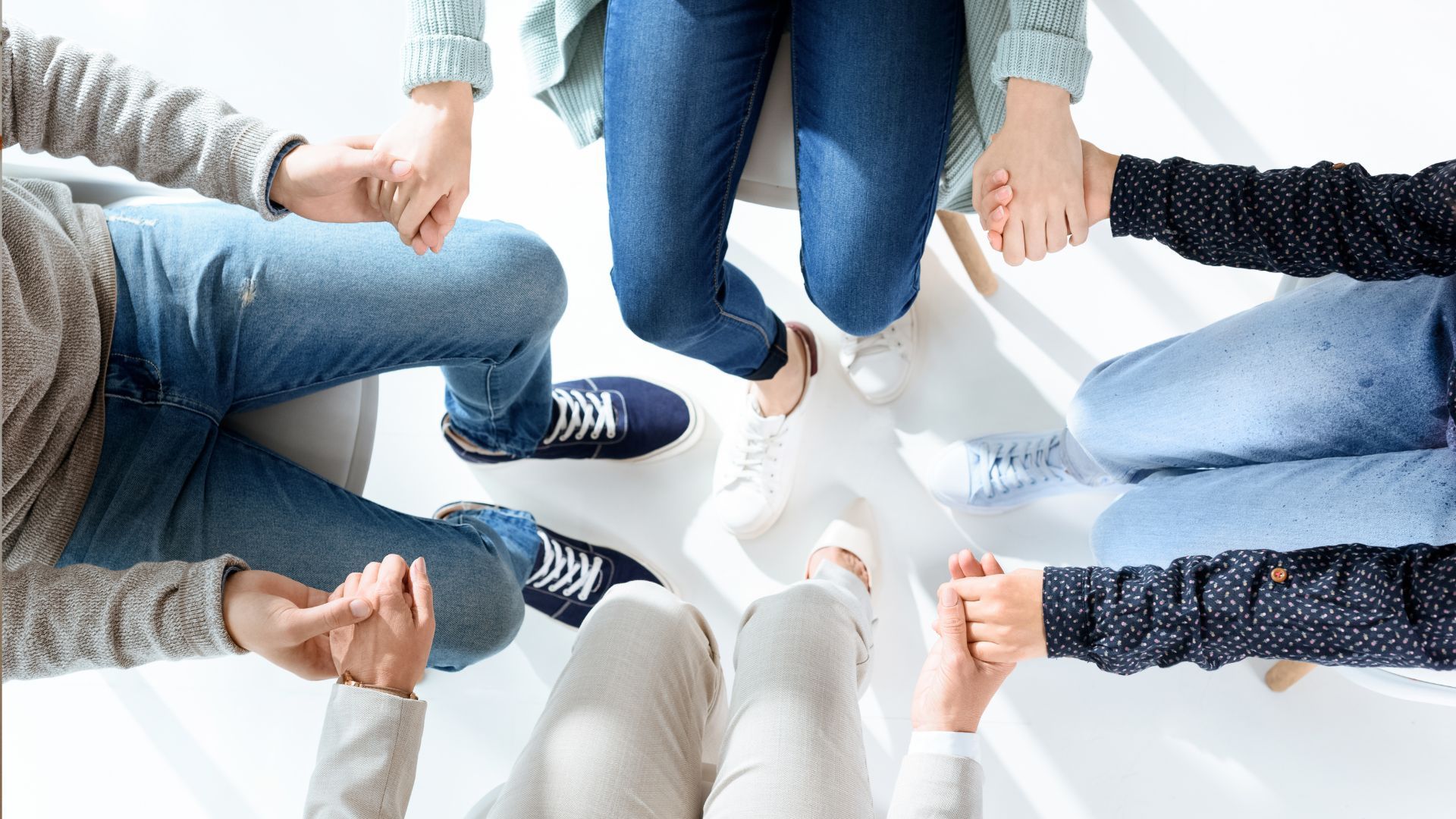 People in a circle, holding hands. Hands visible with legs and feet wearing jeans, shoes, in a well-lit setting.