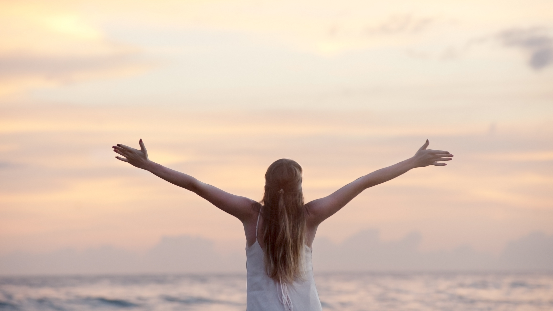 Woman with arms outstretched facing the ocean at sunset.