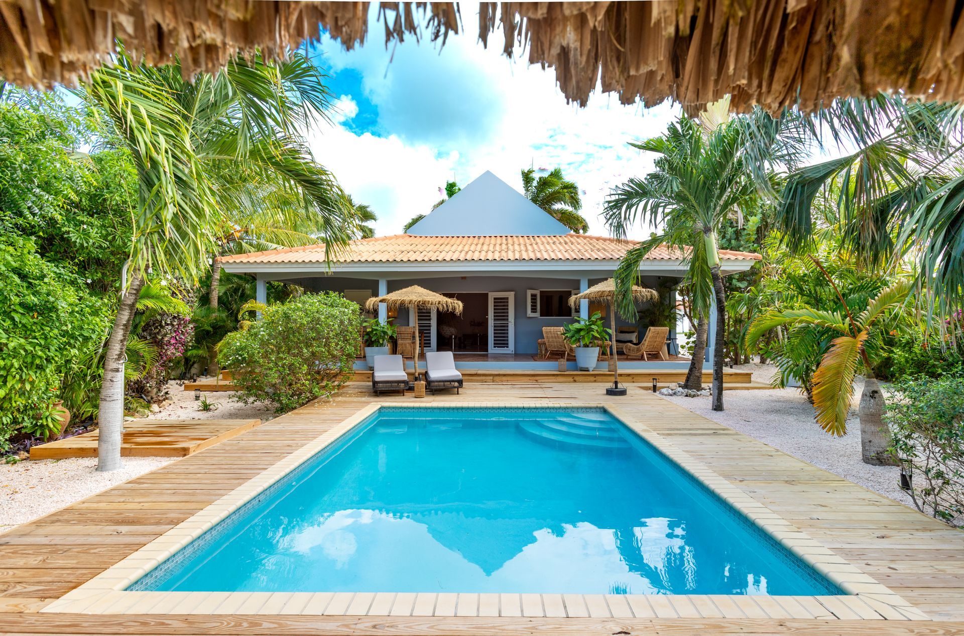 Zwembad voor een huis, palmbomen, blauw water, blauwe lucht en ligstoelen.
