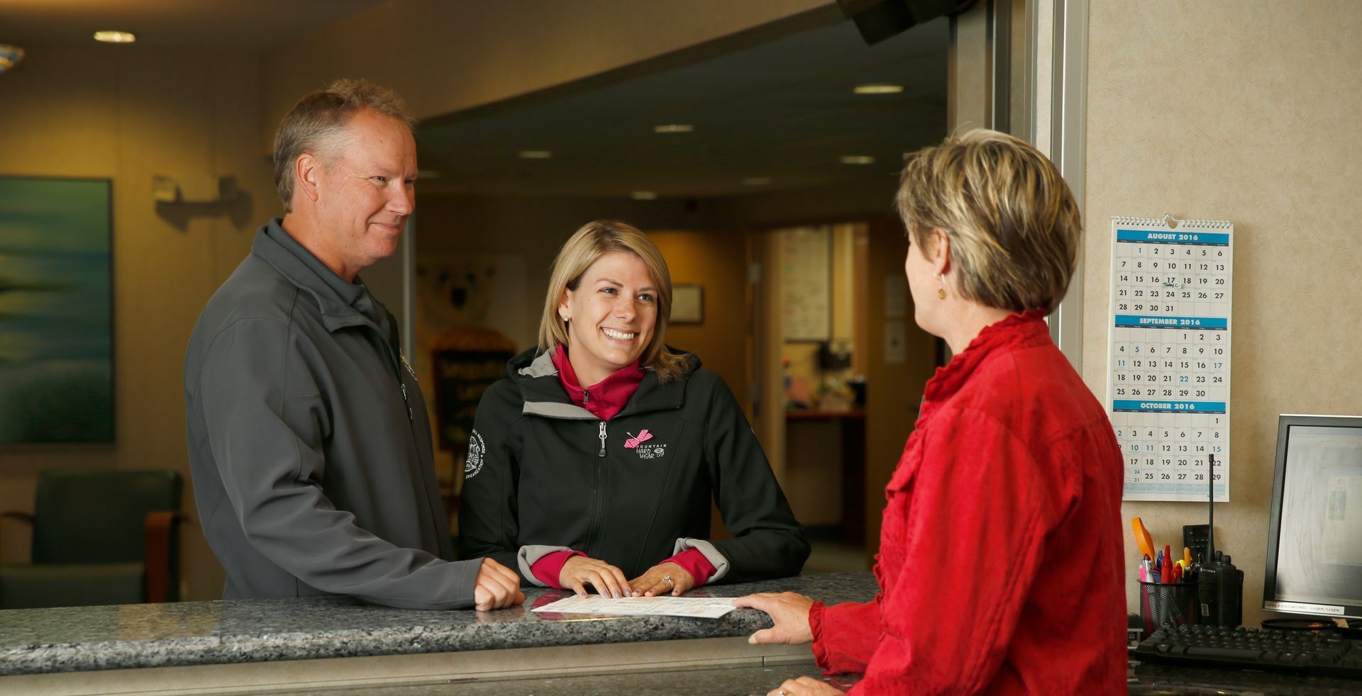 A man and a woman are standing at a counter talking to a woman in a red jacket.