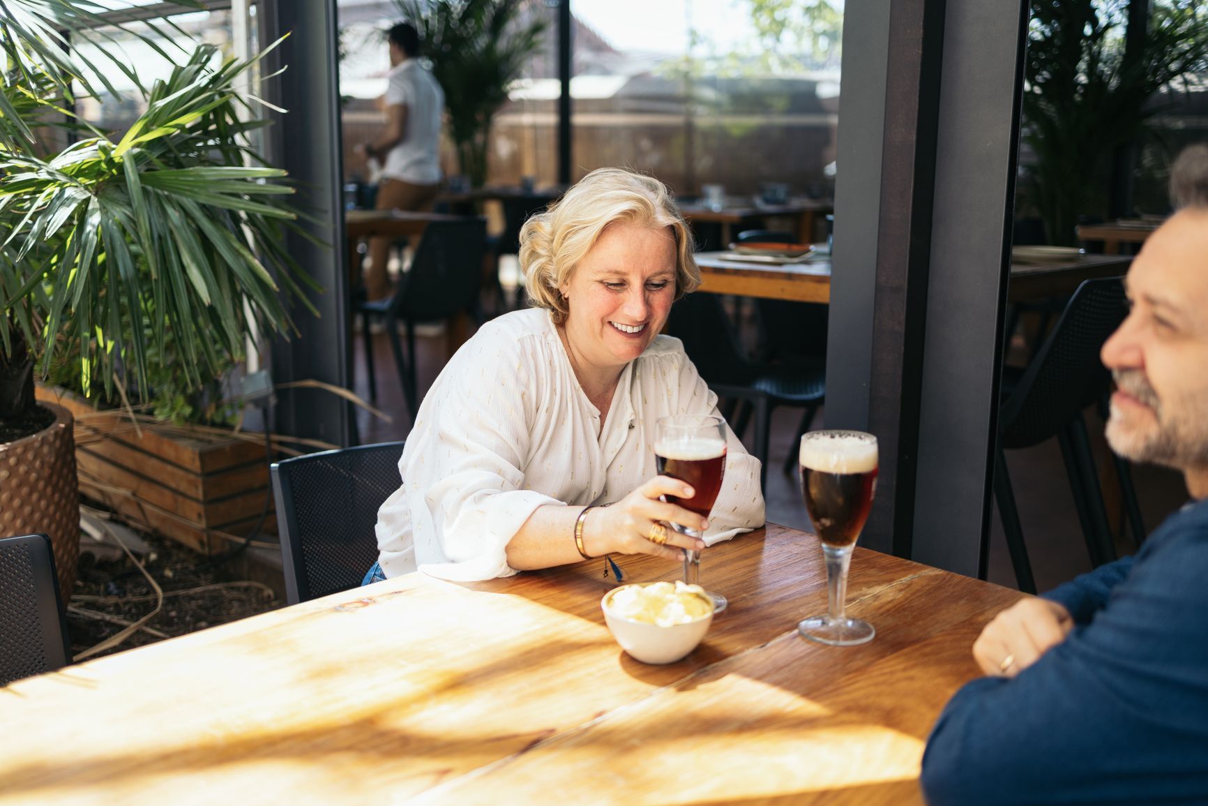 Woman and man at a restaurant table, smiling and holding drinks. Chips in a bowl. Outdoors setting.