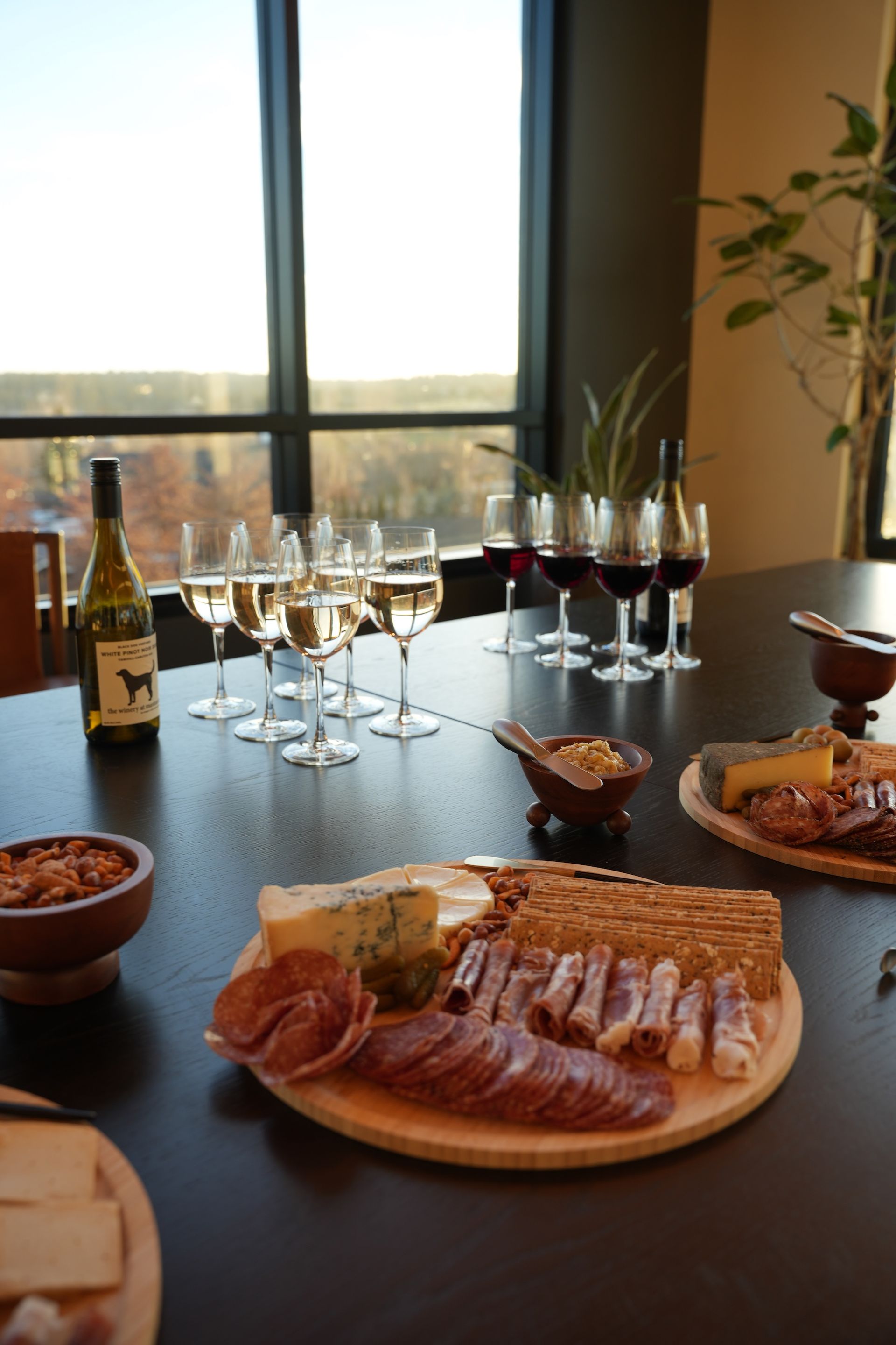 A table with charcuterie boards, bowls of nuts, and wine glasses filled with white and red wine near a large window.